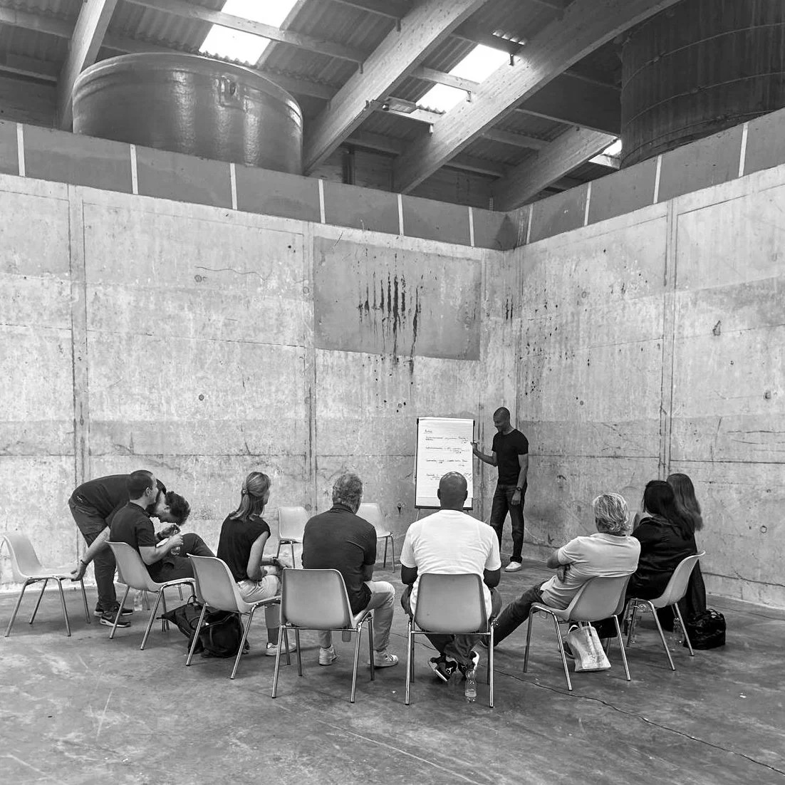 Een groepsfoto van mensen die naar een spreker luisteren in een onaangeklede, industriële ruimte met betonmuren en een houten plafond. Mental health, coaching, welzijn, natuur, kalmte, groei, innerlijke rust, spreker, motivator, coaching, trainer