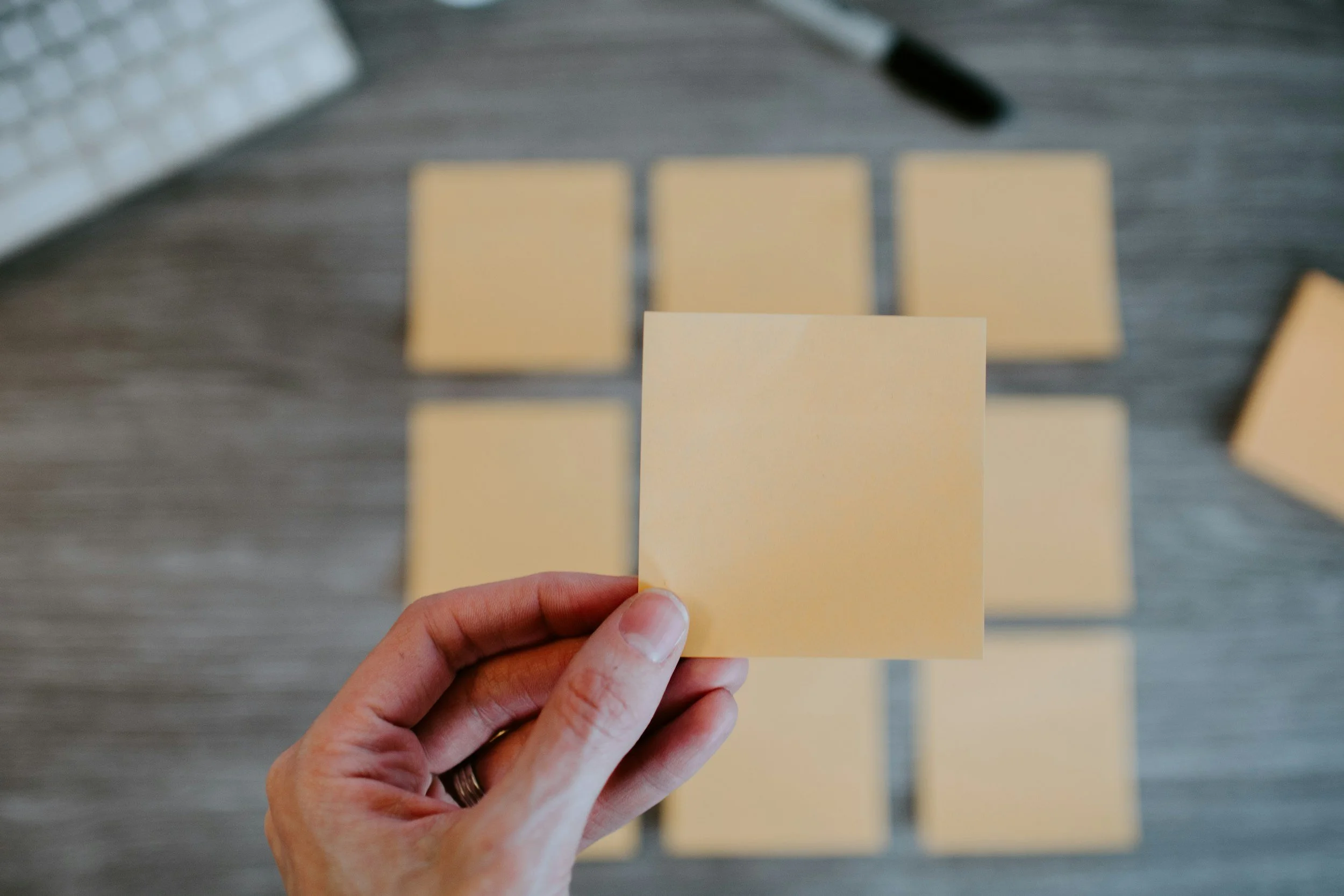 Une main tient un post-it jaune, devant une rangée de plusieurs autres post-its jaunes placés sur une surface en bois, avec un clavier d'ordinateur en arrière-plan.