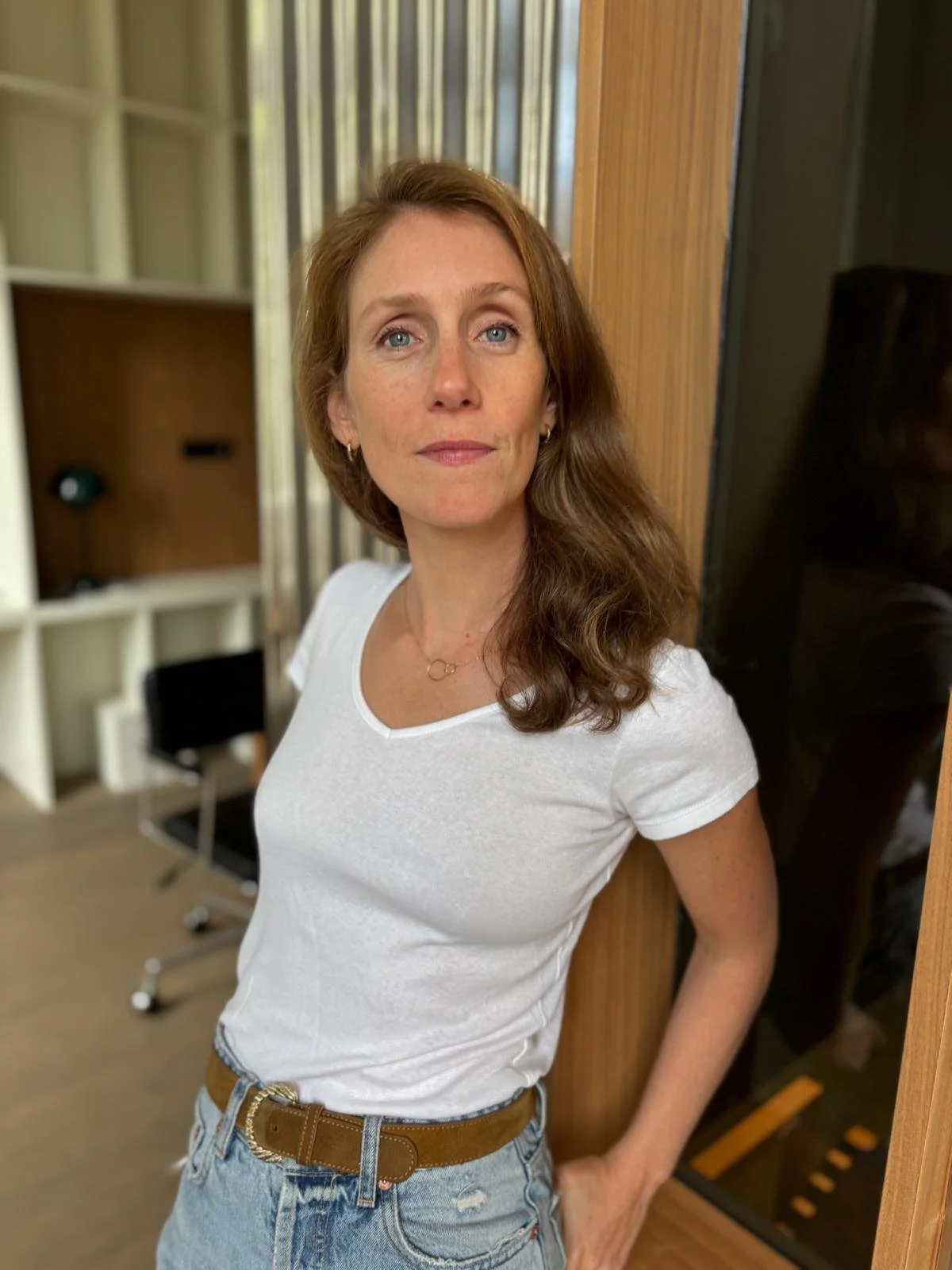 Une femme aux cheveux bruns, portant un t-shirt blanc et un jean décontracté, posant à côté d'une cloison en bois dans un intérieur moderne.