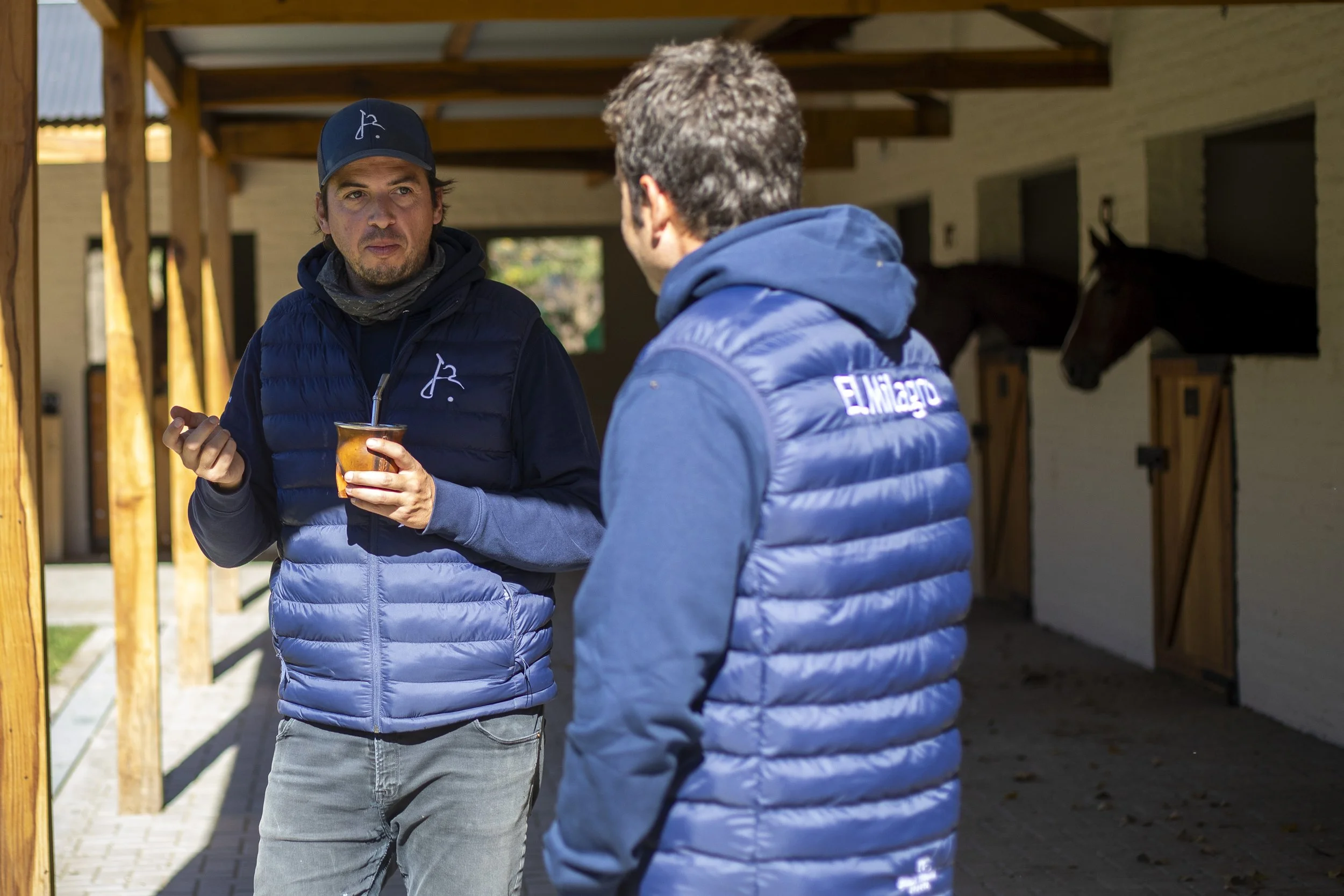 Dos hombres conversan afuera de un establo de caballos, uno sostiene una taza con bebida caliente.