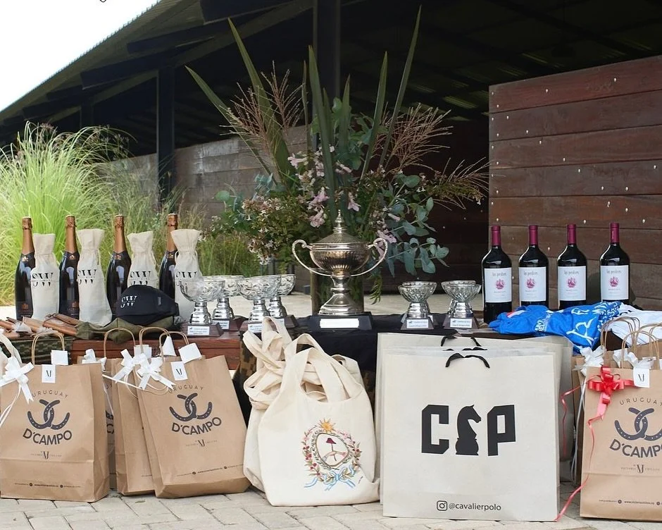 Mesa con varias bebidas como champán y vino, premios en trofeos y kilos, bolsas de regalos de Uruguay D'Campo, y una gran planta decorativa en el centro.