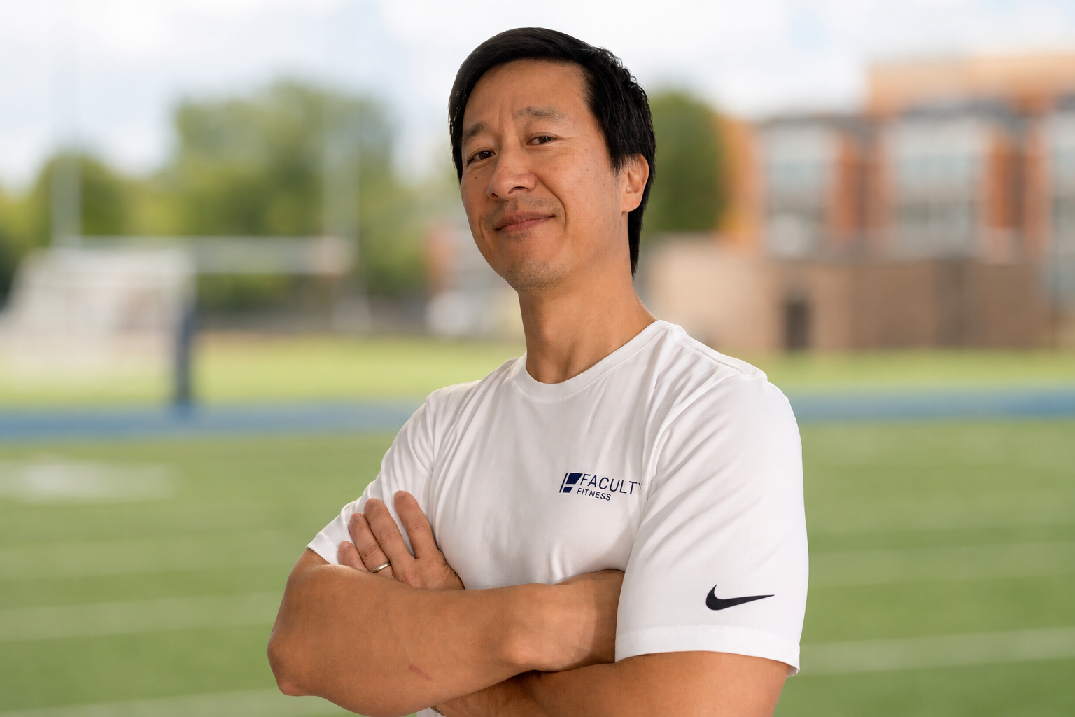 Man in white athletic shirt with 'Faculty Fitness' logo, arms crossed, standing outdoors on a sports field with blurred green grass and trees in the background.