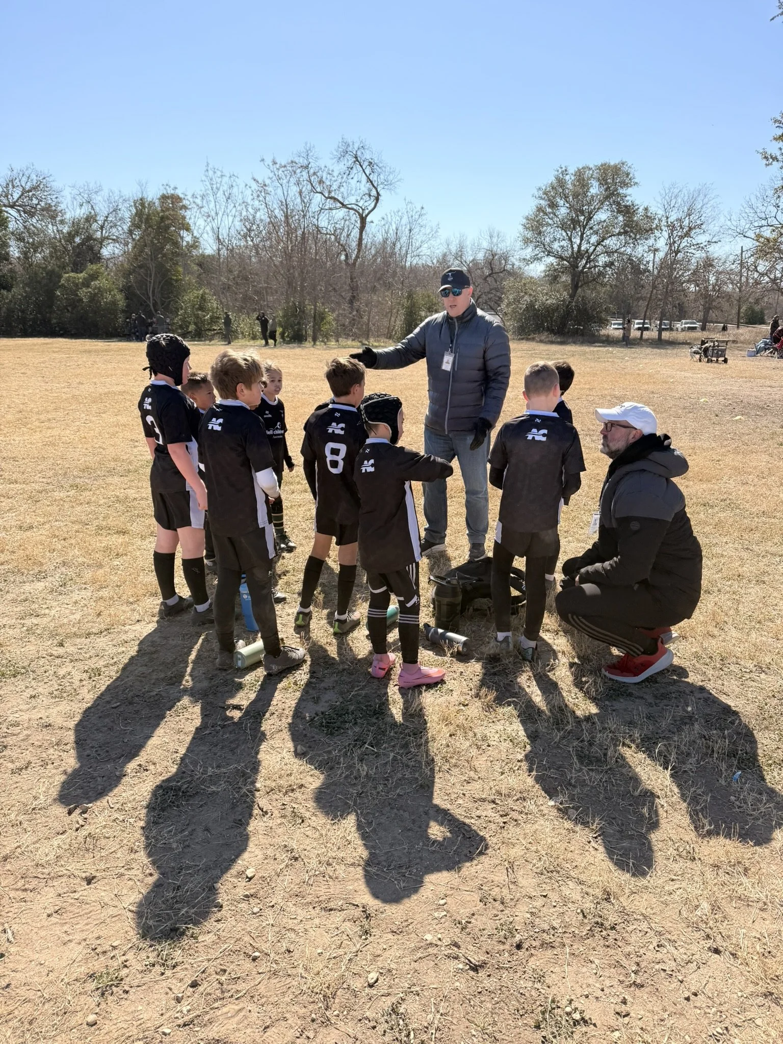 Adult coach speaking to young children rugby players on an outdoor field with dry grass and leafless trees in the background on a sunny day.