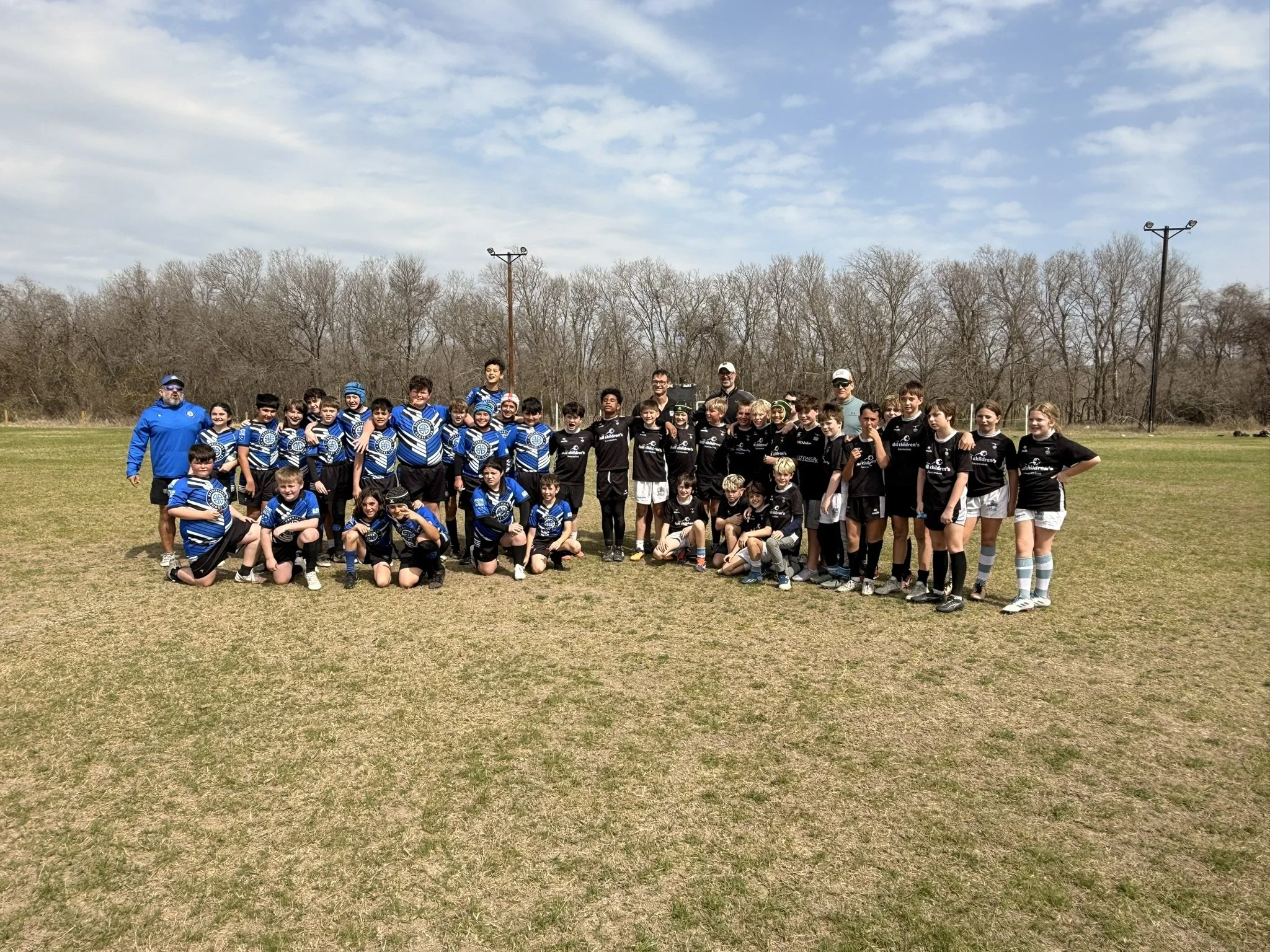 A group of children and adults posing for a photo on a grassy field, with leafless trees and a cloudy sky in the background.