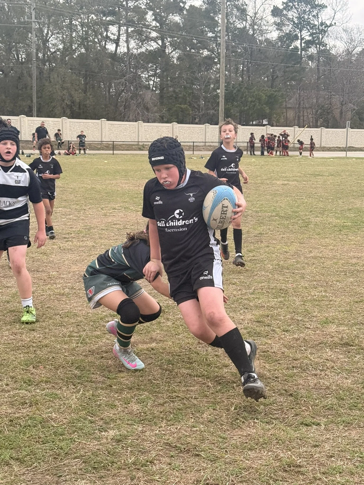 Young rugby players in black and white jerseys on a grassy field during a game, with some players running and others on the ground, and a white fence and trees in the background.