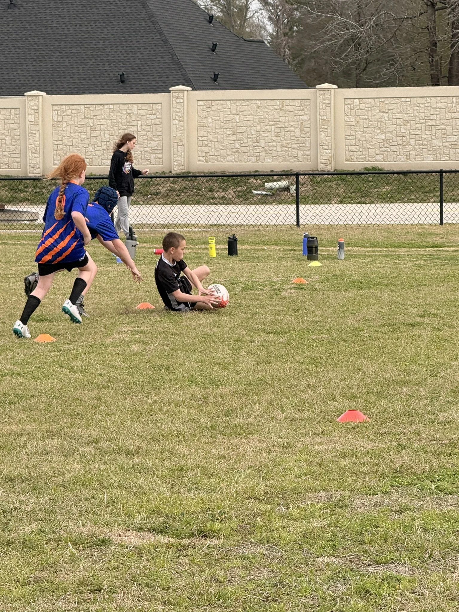 A group of children playing rugby on a grassy field enclosed by a black chain-link fence, with a beige stone wall and a house with a dark gray roof in the background.