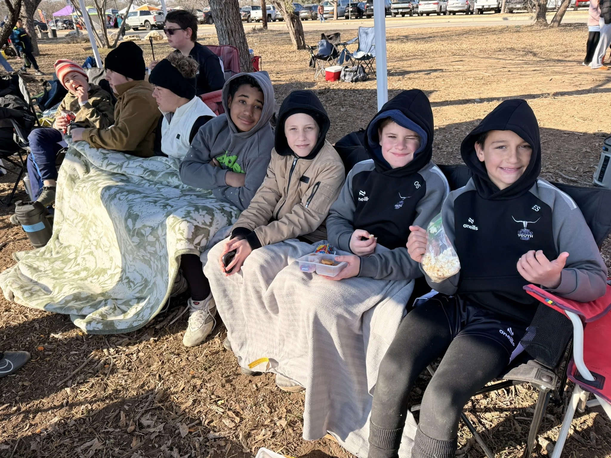 Group of boys sitting on a bench outdoors between games, some eating popcorn and snacks, wearing jackets and hoodies, with trees, parked cars, and people in the background on a sunny day.