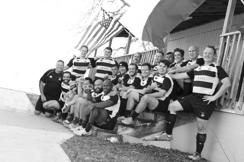 A group of rugby players in black and white striped uniforms sitting and standing together outdoors near a beach, with American flags hanging above them.