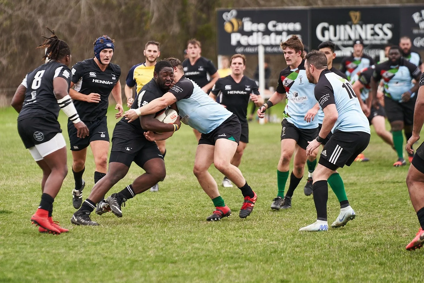 Rugby players in black and light blue jerseys compete for the ball on a grassy field, with several players and a sign in the background.