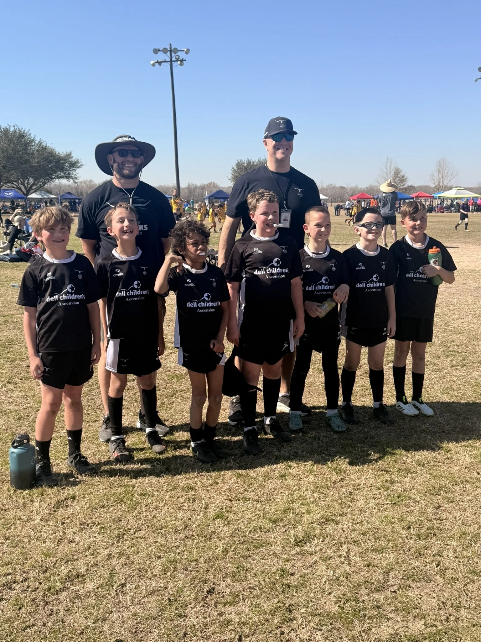 Group of young boys in black sports uniforms with sponsors' logos, standing on a grassy field with two adult coaches, at an outdoor sports event under a clear blue sky. Tents and other athletes are visible in the background.