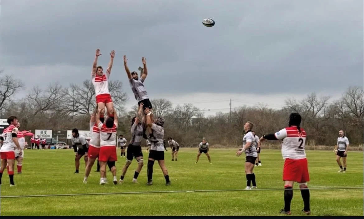 Rugby players in the middle of a line-out, lifting a player to catch the ball during a match on a green field with trees and cloudy sky in the background.