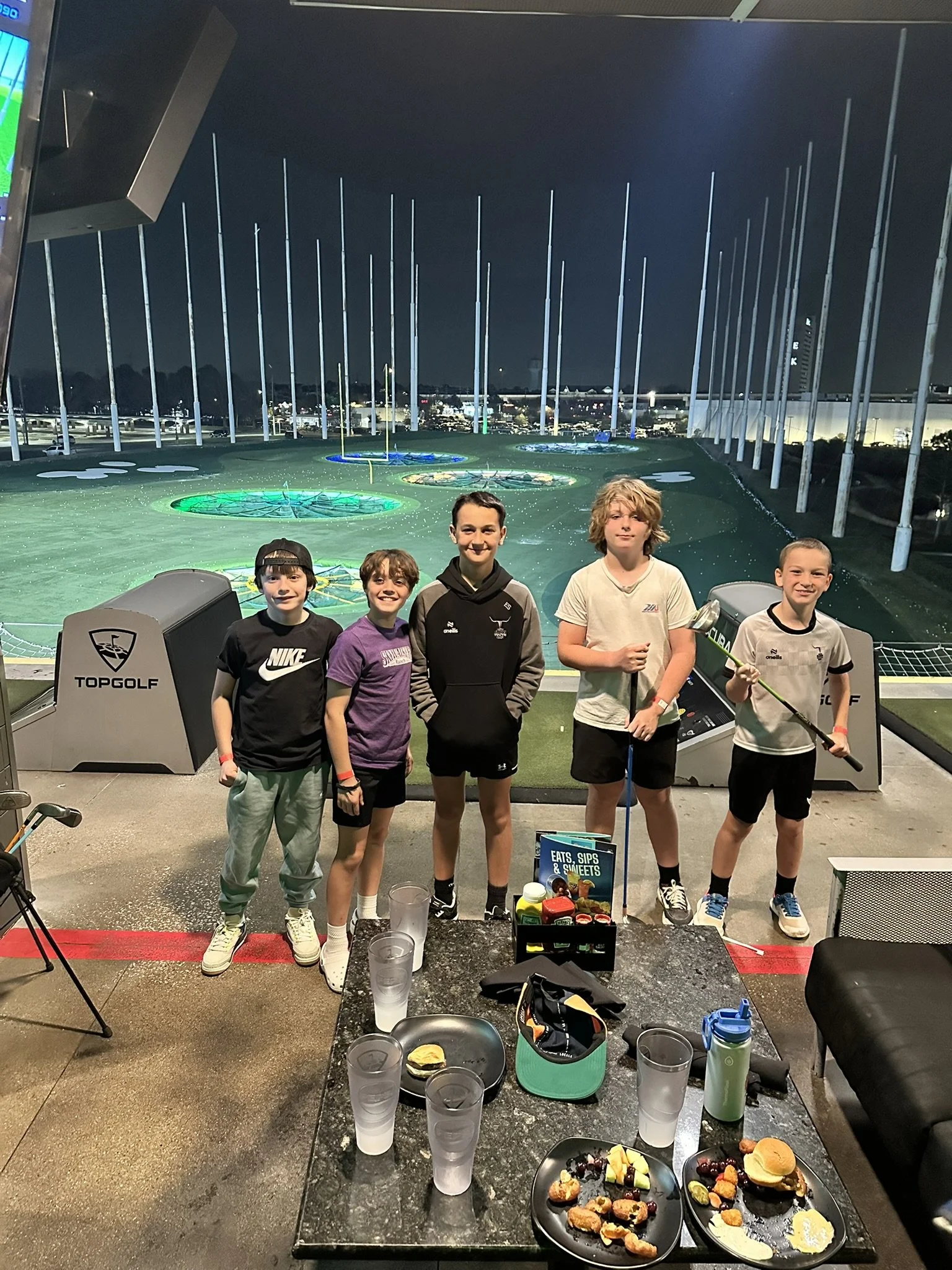 Five children standing together at a team Topgolf outing with plates of food, drinks, and snacks, with a nighttime golf course in the background.