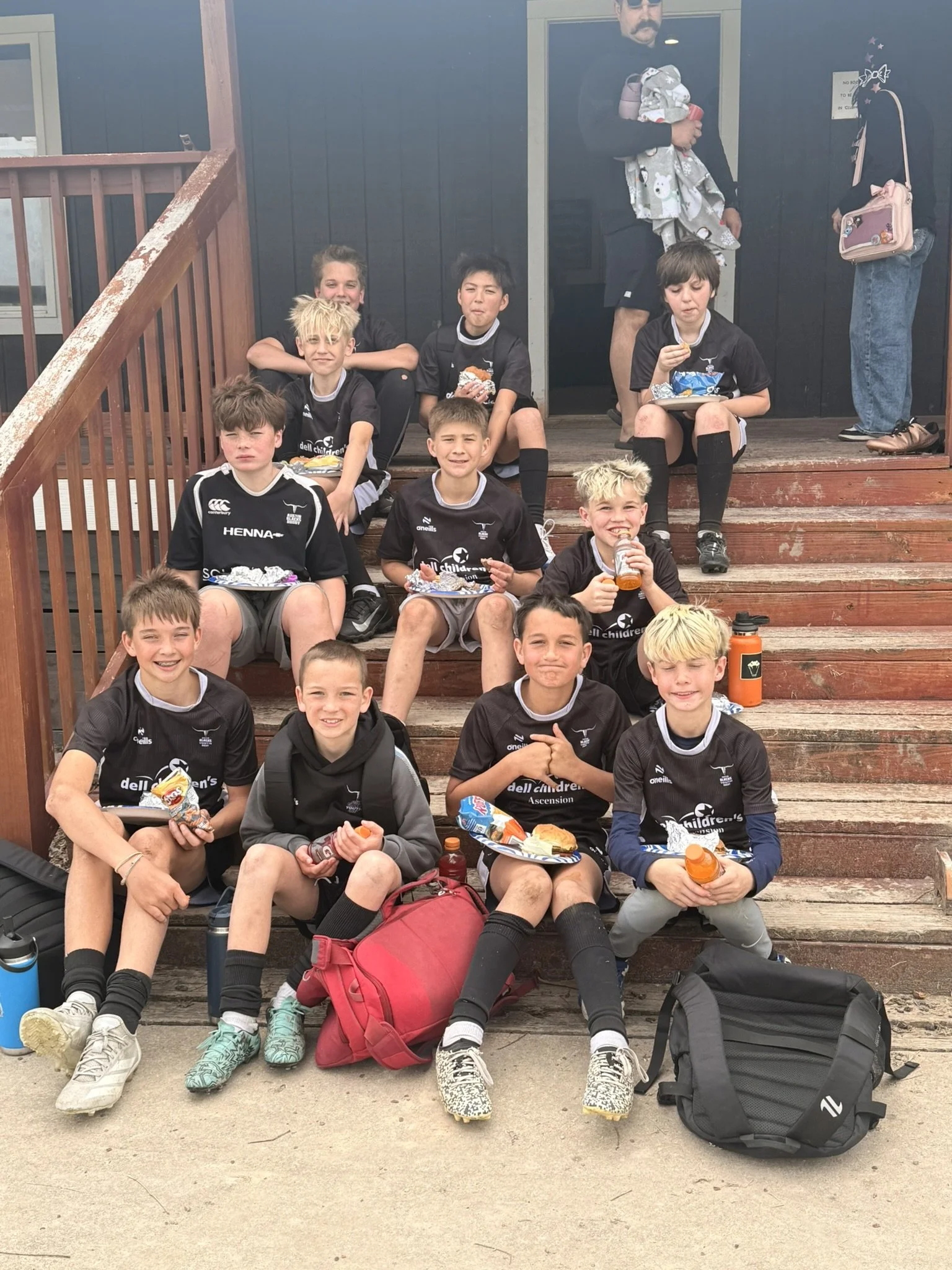 A group of young boys in matching Austin Blacks jerseys, sitting and standing on wooden steps outside the Austin Blacks clubhouse after a friendly match with Katy, enjoying snacks and drinks, with backpacks around them.