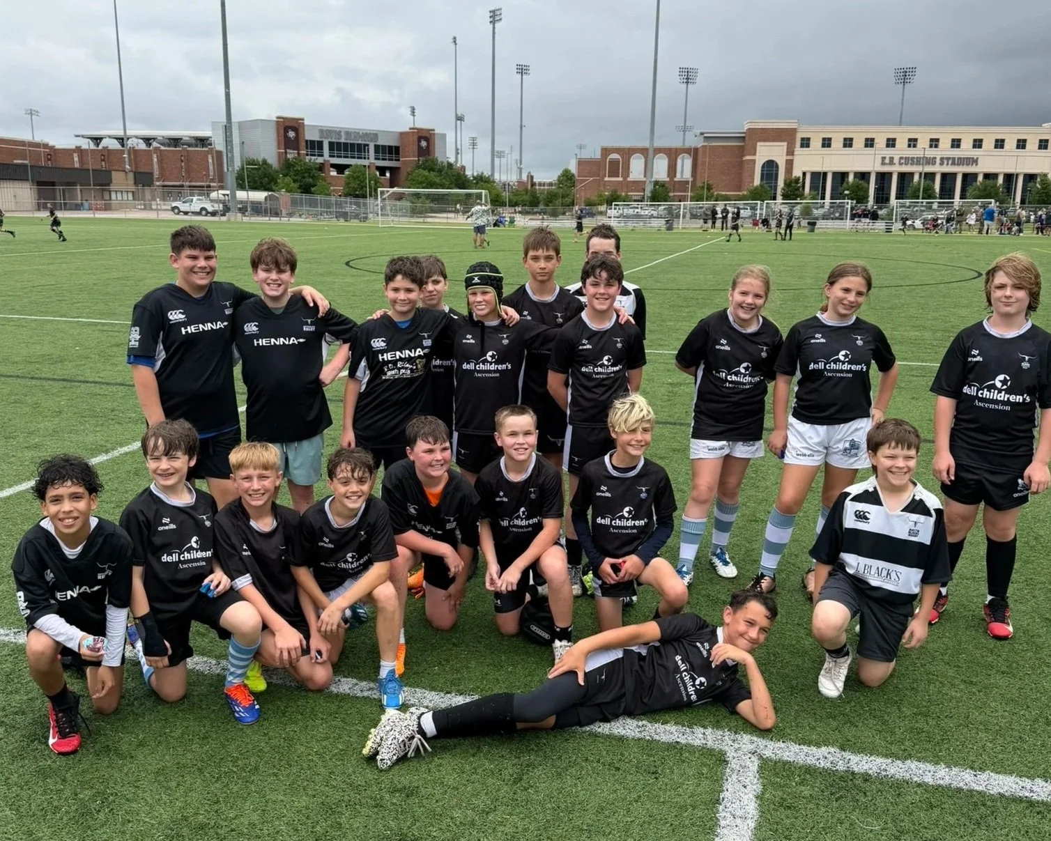 A group of young children and teenagers in black and white rugby uniforms posing on a sports field with a stadium in the background.