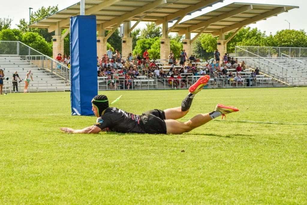 A rugby player falling or diving on the grass field during a game, near the goalpost pad, with a crowd of spectators seated in the stands in the background.