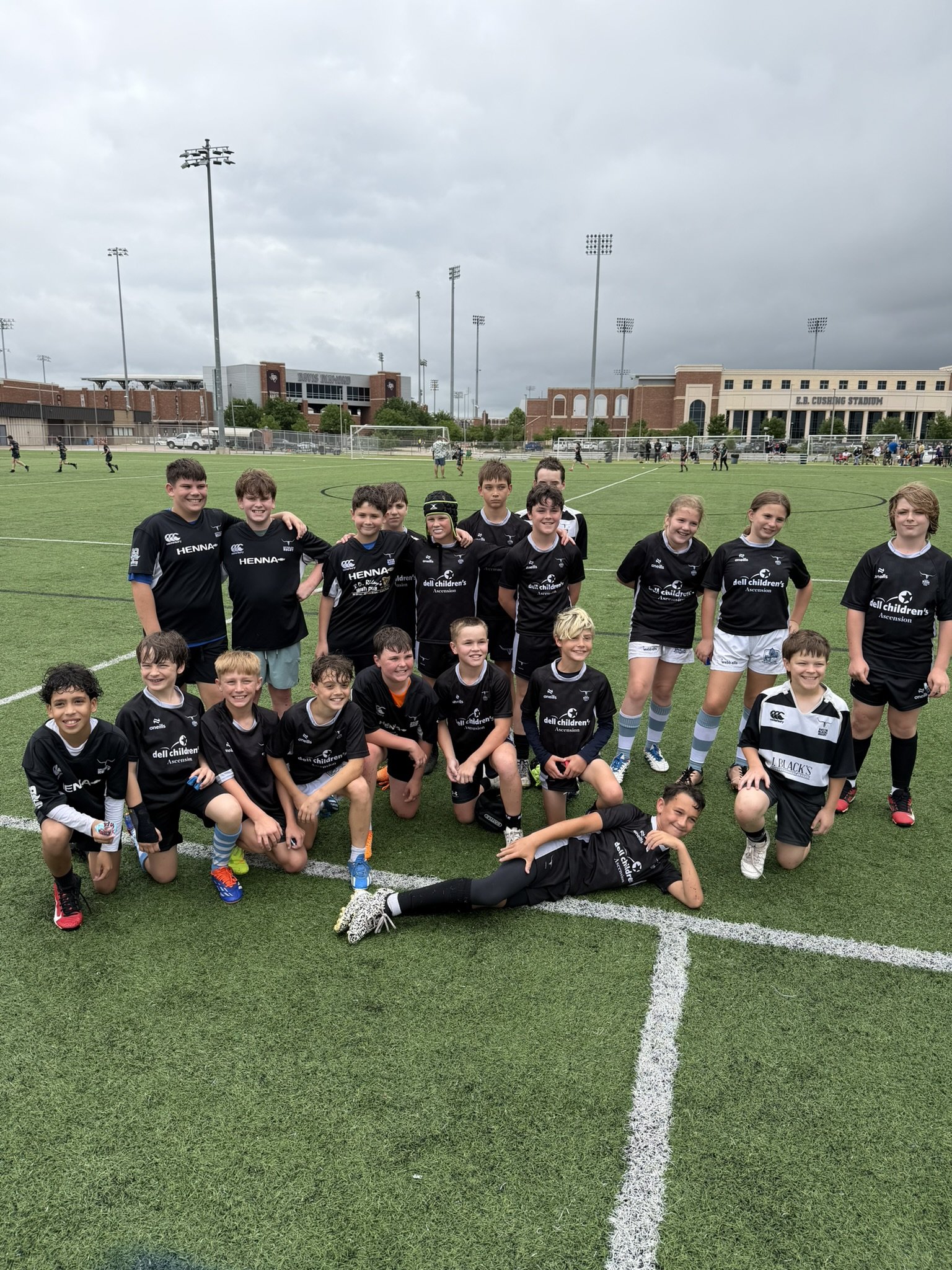 Group of children and some adults on a soccer field, some standing and some kneeling or lying down, with a stadium and cloudy sky in the background.