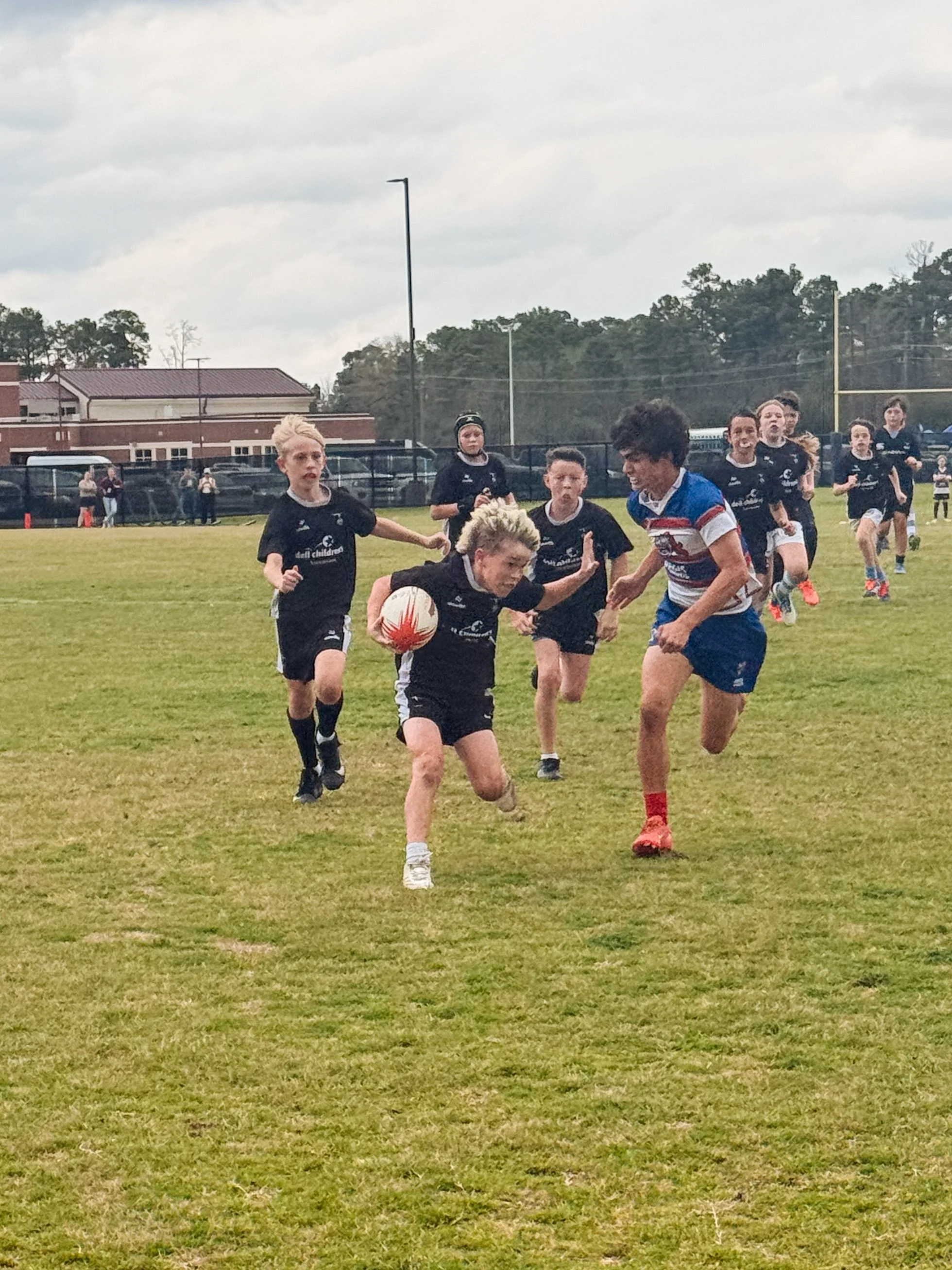 Youth rugby players on a grassy field competing for possession of a rugby ball, with some players in black uniforms and one in a red, white, and blue striped uniform.