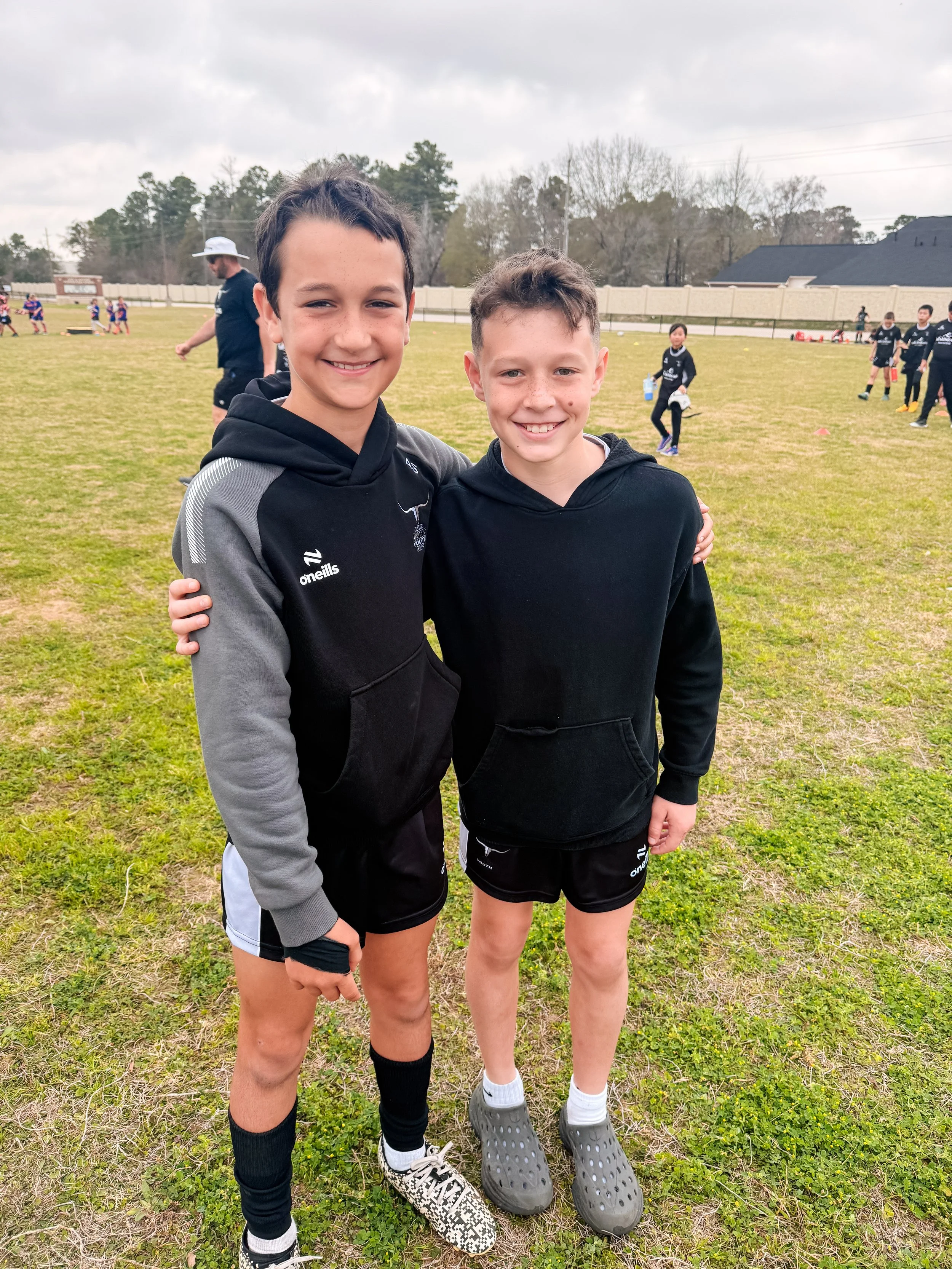 Two young boys standing outdoors on a grassy field, smiling at the camera; one is wearing a black and gray athletic hoodie, shorts, and patterned sneakers, the other is in a black hoodie, shorts, and slip-on shoes; in the background, there are other children and a coach, with a tennis court and trees behind them.