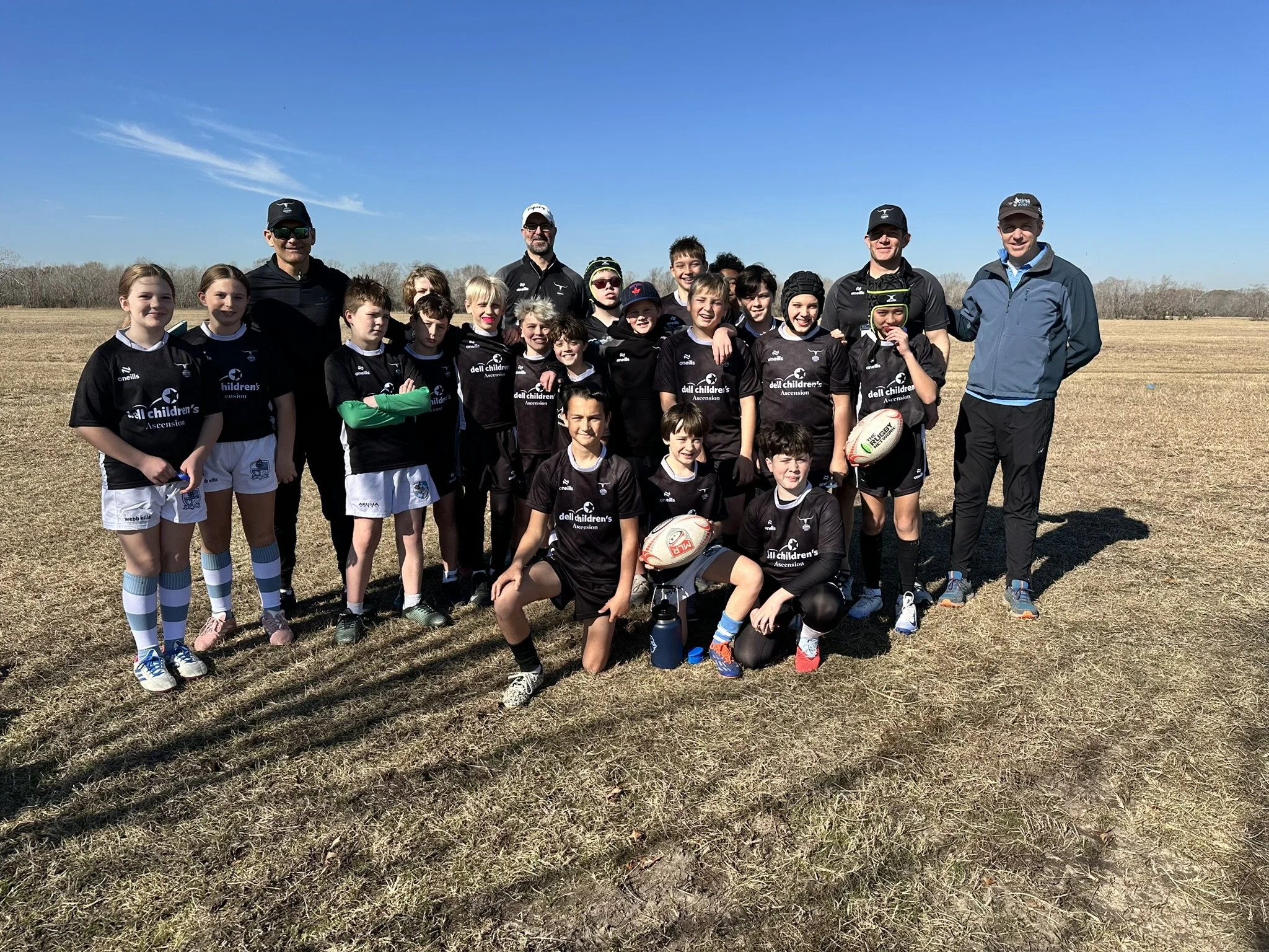 A group of children and adults, some in sports uniforms, standing on a dry grassy field under a clear blue sky, posing for a team photo.