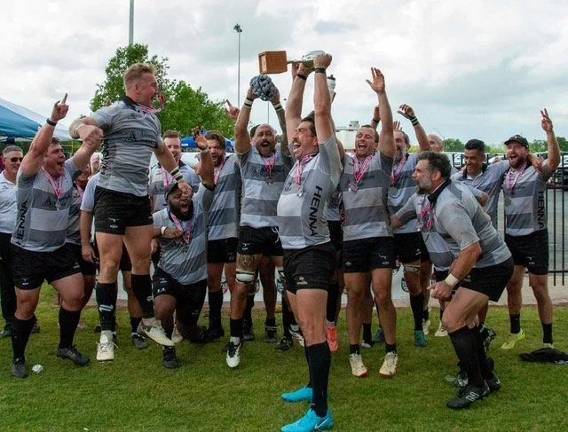 A rugby team celebrating on the field, with one player lifting a trophy, surrounded by teammates cheering and raising their hands.