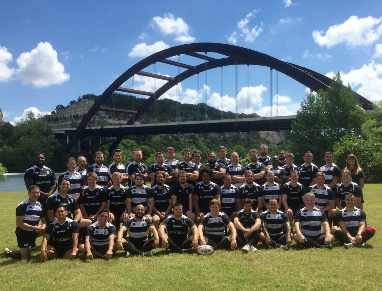 Group photo of rugby players in black and white jerseys, sitting and standing on grass field under the Austin Pennybacker bridge with hills and clouds in the background.