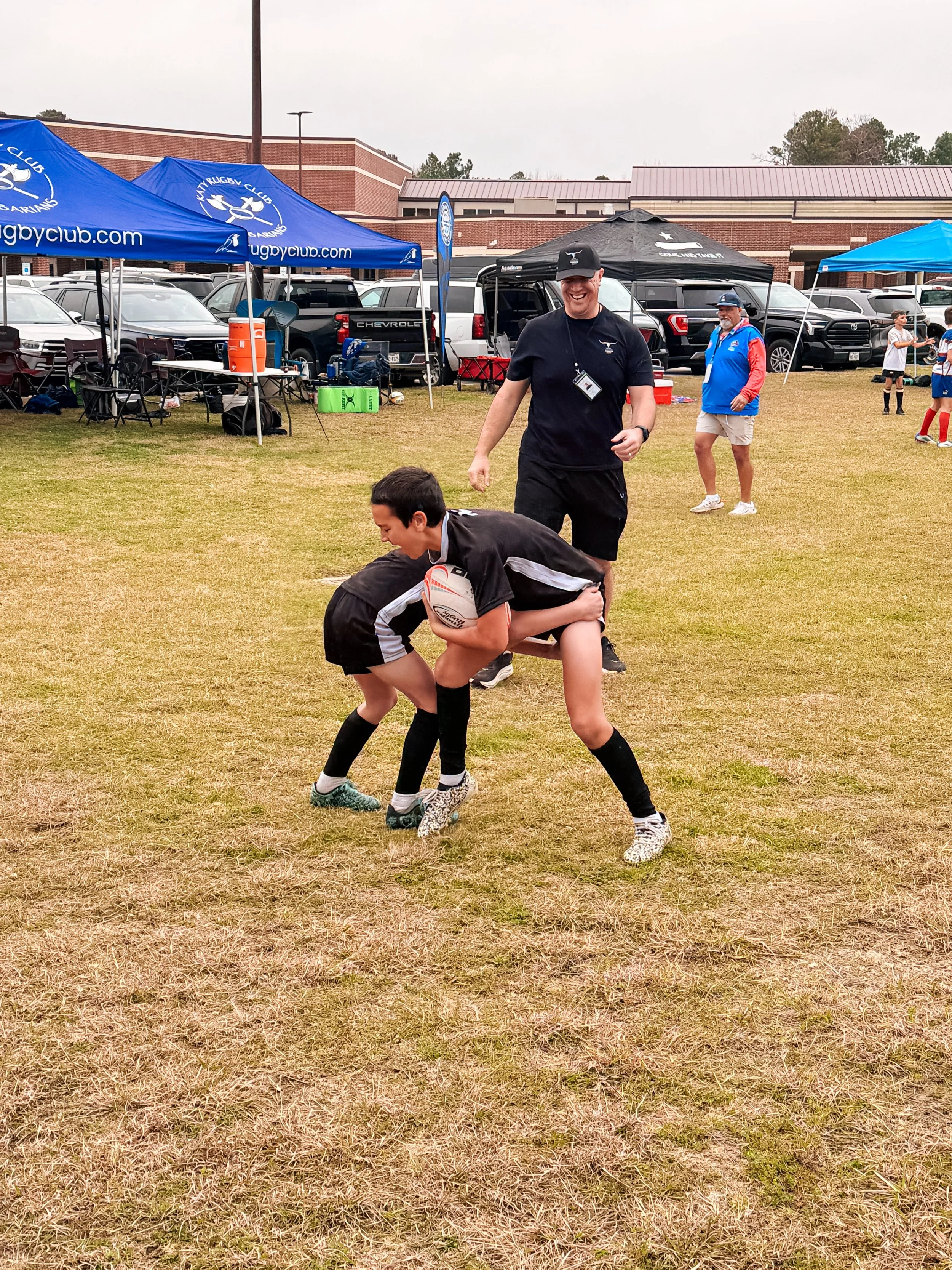 A youth rugby game on a grassy field with coaches and spectators under tents, some parked cars, and a brick building in the background. The game involves a young player holding a rugby ball, being tackled by another player, while a coach smiles nearb