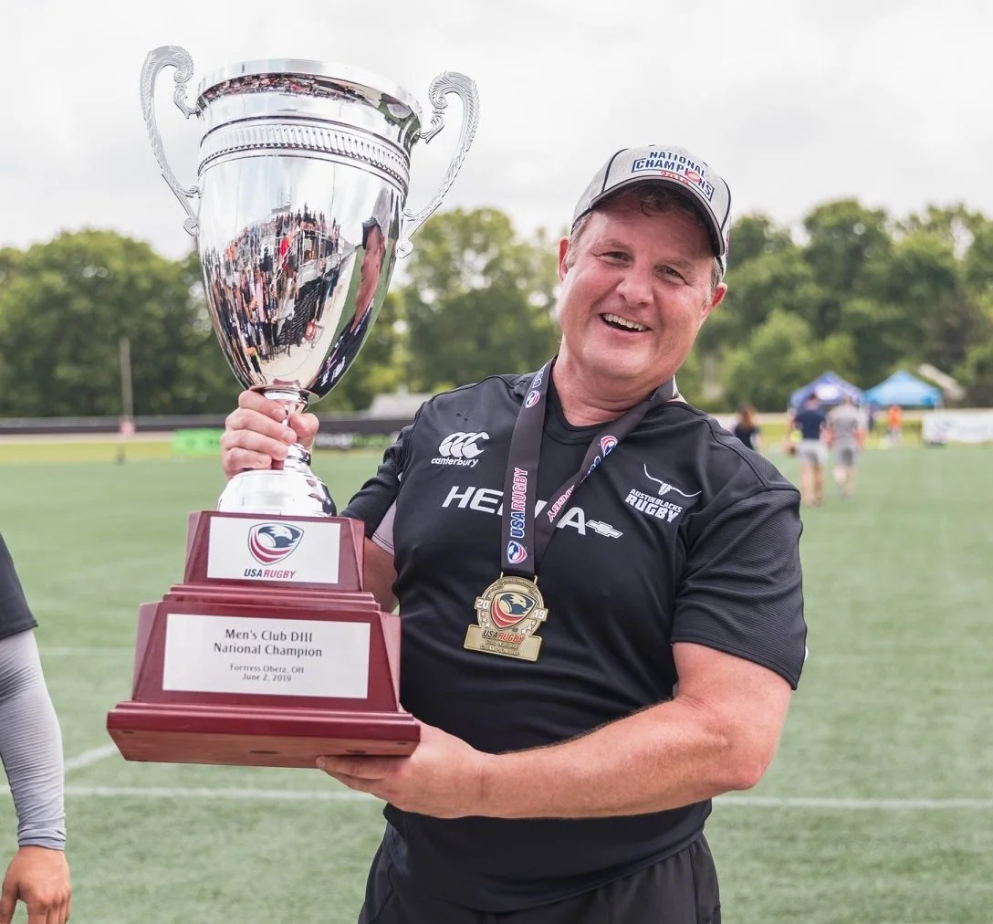A man smiling and holding a large silver trophy while wearing a medal around his neck, at a sports field during daytime.