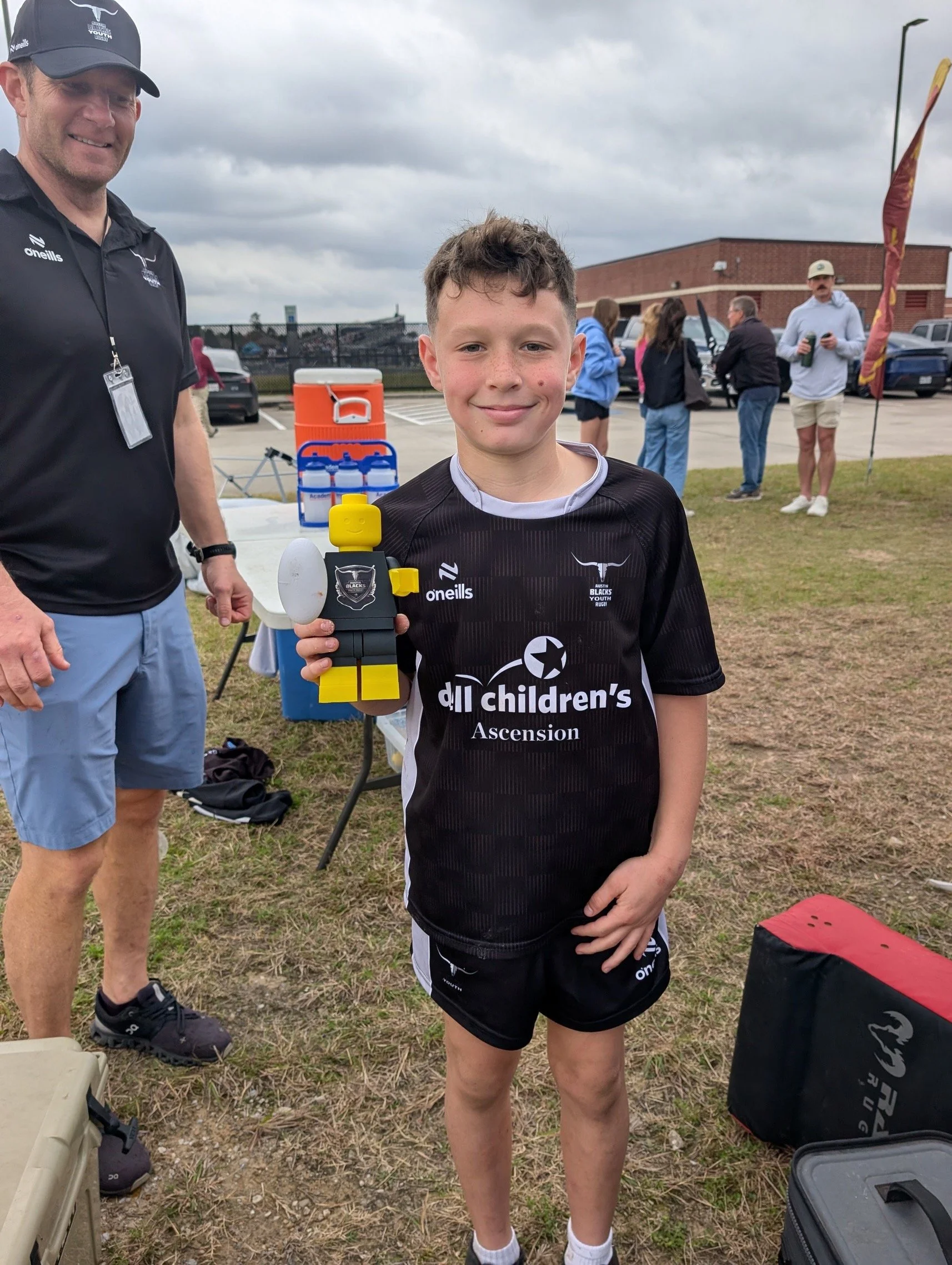 Young boy holding a LEGO trophy with a smile, dressed in black sportswear, standing outdoors with a group of people and a building in the background.