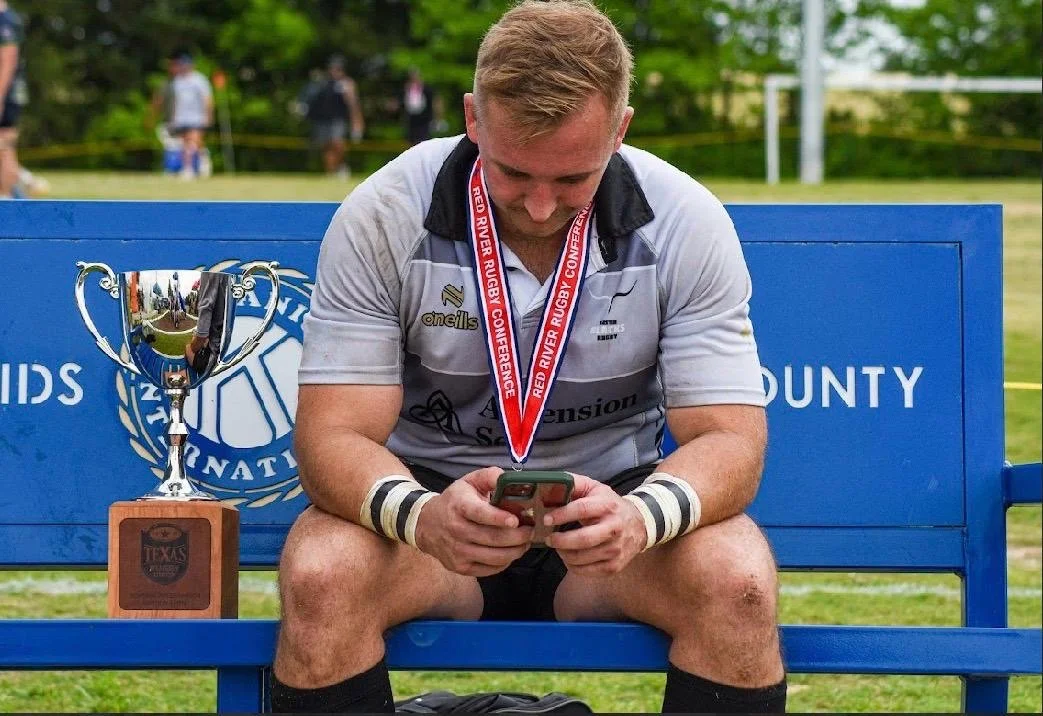 A young male rugby player sitting on a blue bench, looking at his phone, wearing a medal around his neck, with a trophy and a blue sign behind him.
