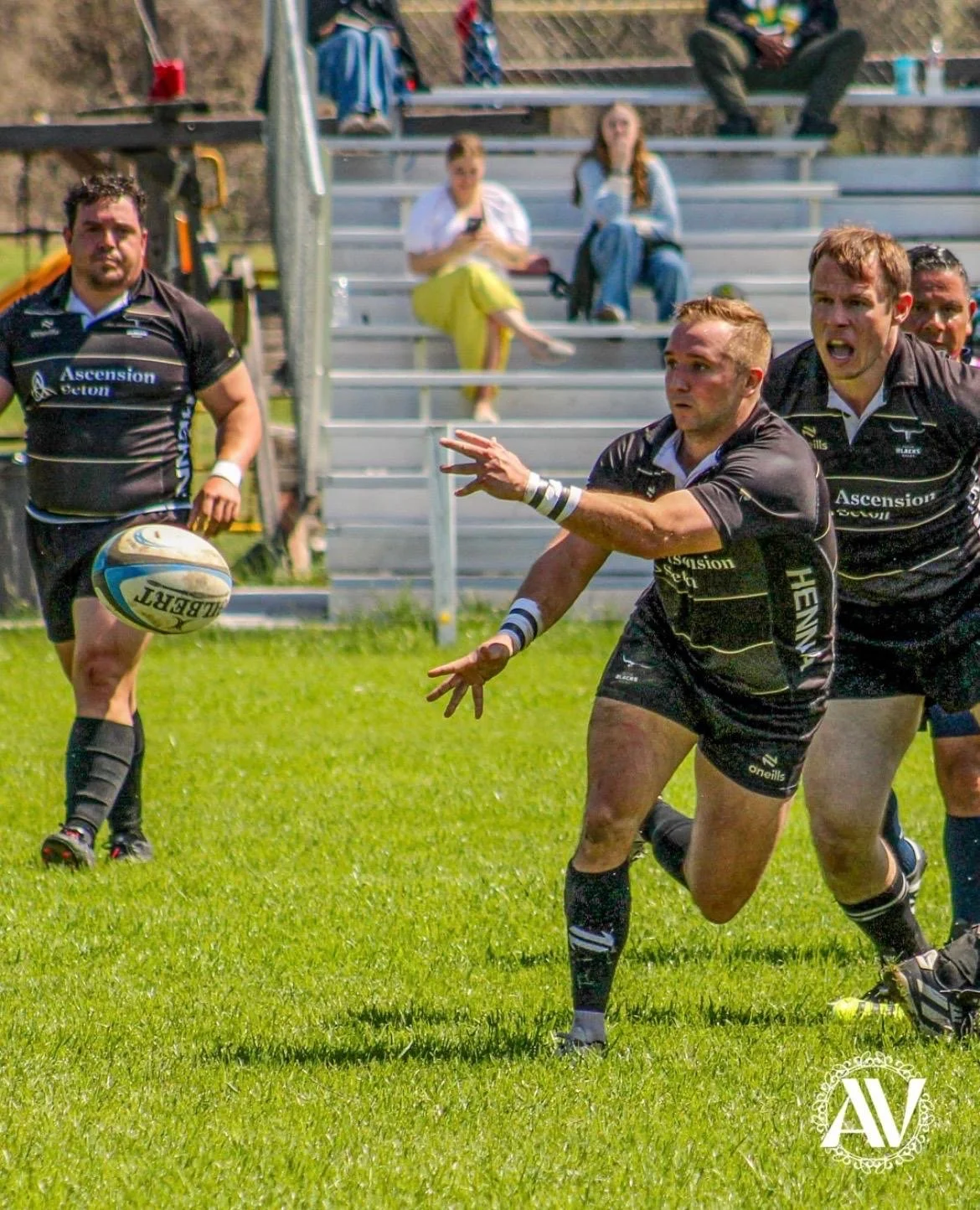 Rugby players in black uniforms on a grassy field, with one player passing the ball and others running behind him, spectators sitting on bleachers in the background.