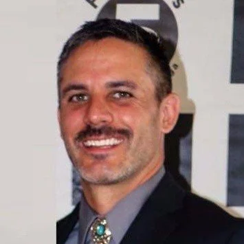 A man with short dark hair and a mustache, smiling, wearing a suit jacket and a gray shirt, standing in front of a background with text and logos.