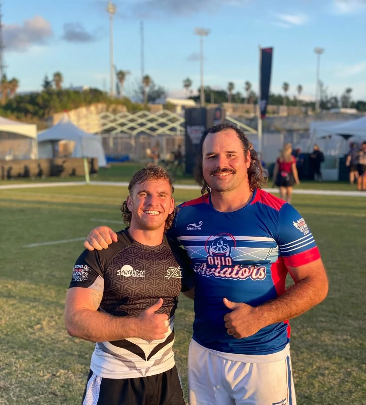 Two men standing on a rugby field, smiling and giving thumbs up. One wears a black and white rugby jersey, the other a red, white, and blue jersey with 'Ohio Guardians' written on it. Tents and other people are in the background.