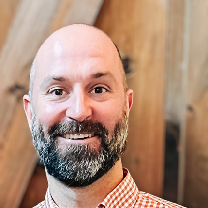 Close-up of a smiling man with a beard and shaved head, wearing a checkered shirt, in front of a wooden background.
