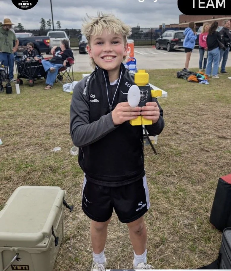 A young boy with blonde hair smiling and holding a yellow and black Lego robot at an outdoor sports event, with other people and parked cars in the background.