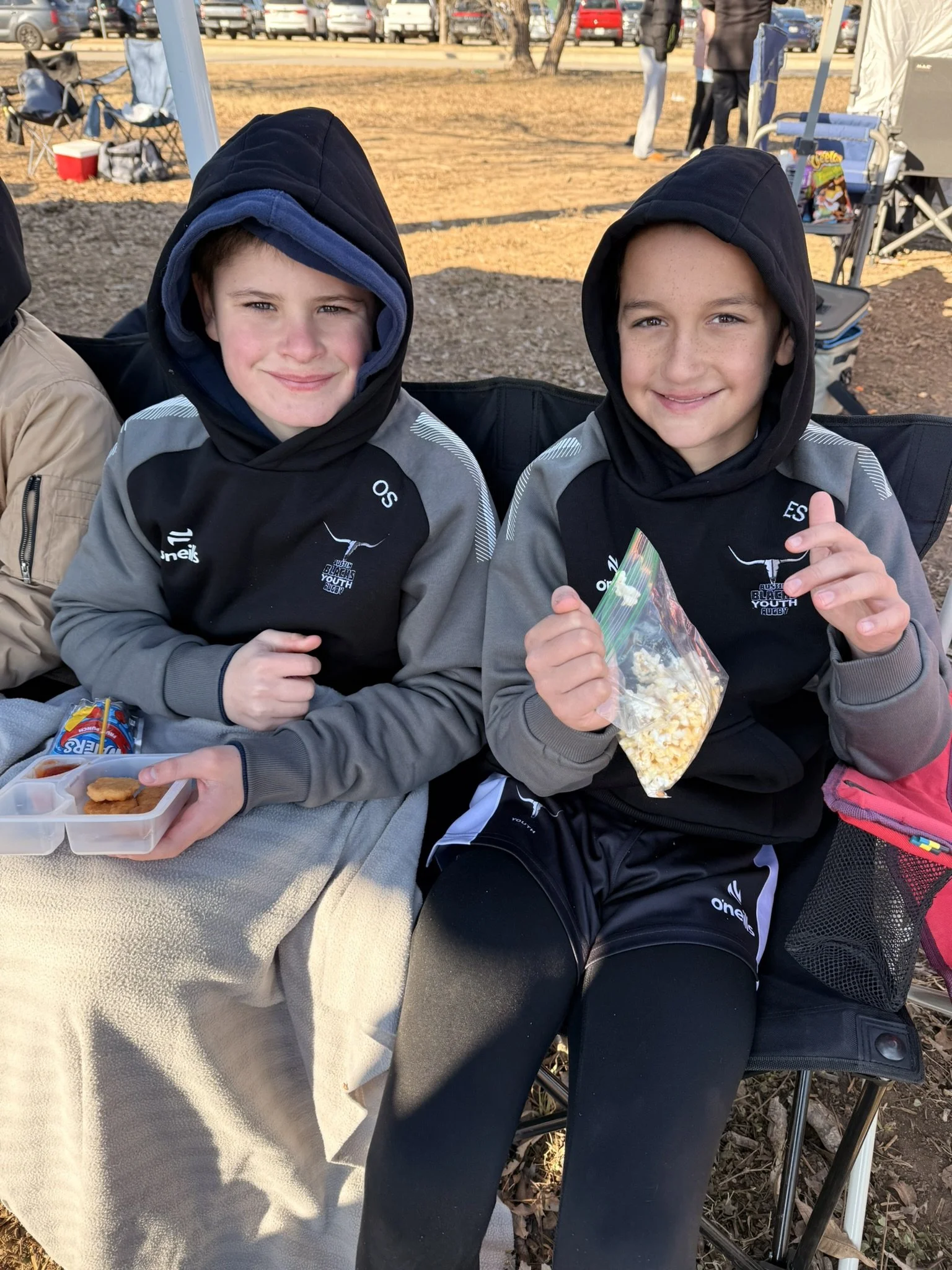 Two boys in black hoodies with gray accents and sports logos sitting at a table outdoors, smiling at the camera, holding snacks, with fallen leaves and a parking lot in the background.