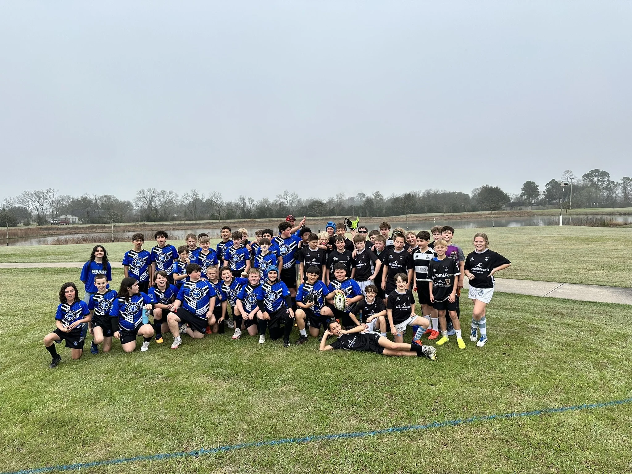 A large group of children and teens wearing sports uniforms, gathered together on a field for a team photo, with a lake and trees in the background on an overcast day.