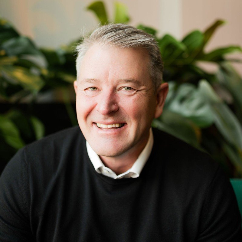 A smiling middle-aged man with gray hair, wearing a black sweater over a white collared shirt, sitting indoors in front of green plants.