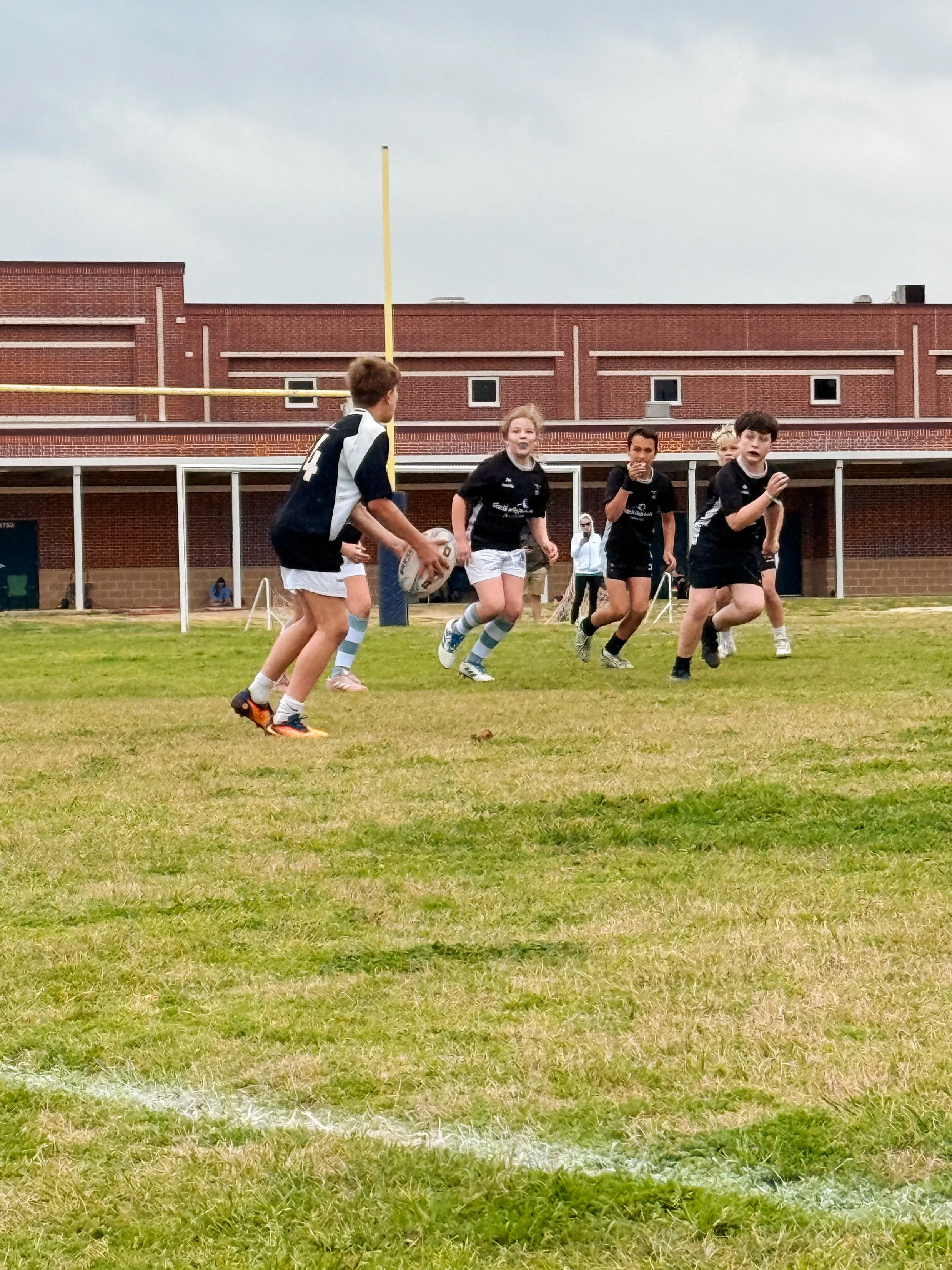 Children playing rugby on a grassy field in front of a red brick building, with overcast skies.