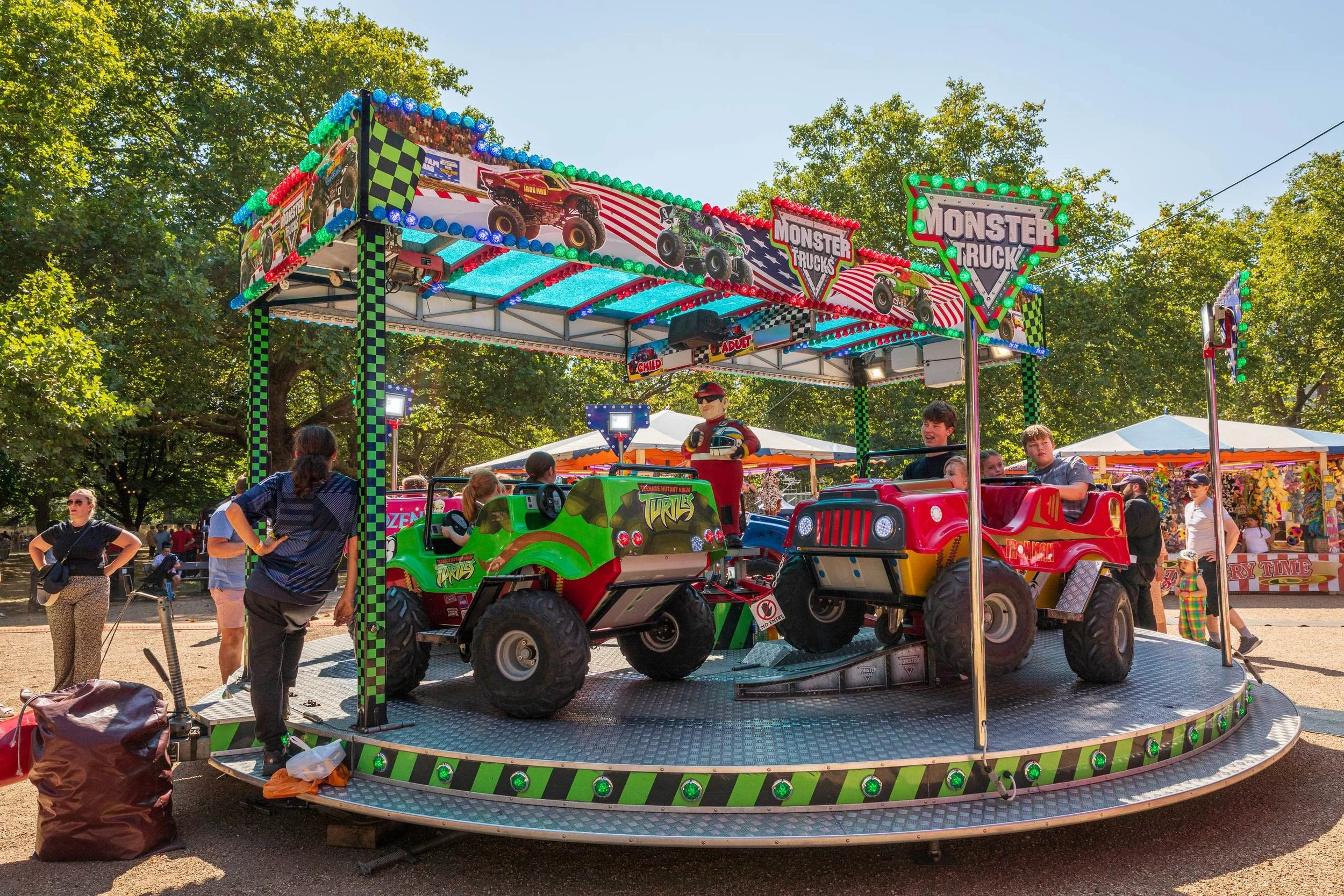 Children and adults at an amusement park riding a Kid's Monster Truck ride with toy monster trucks and a sign labeled 'Monster Trucks' outdoors during daytime.