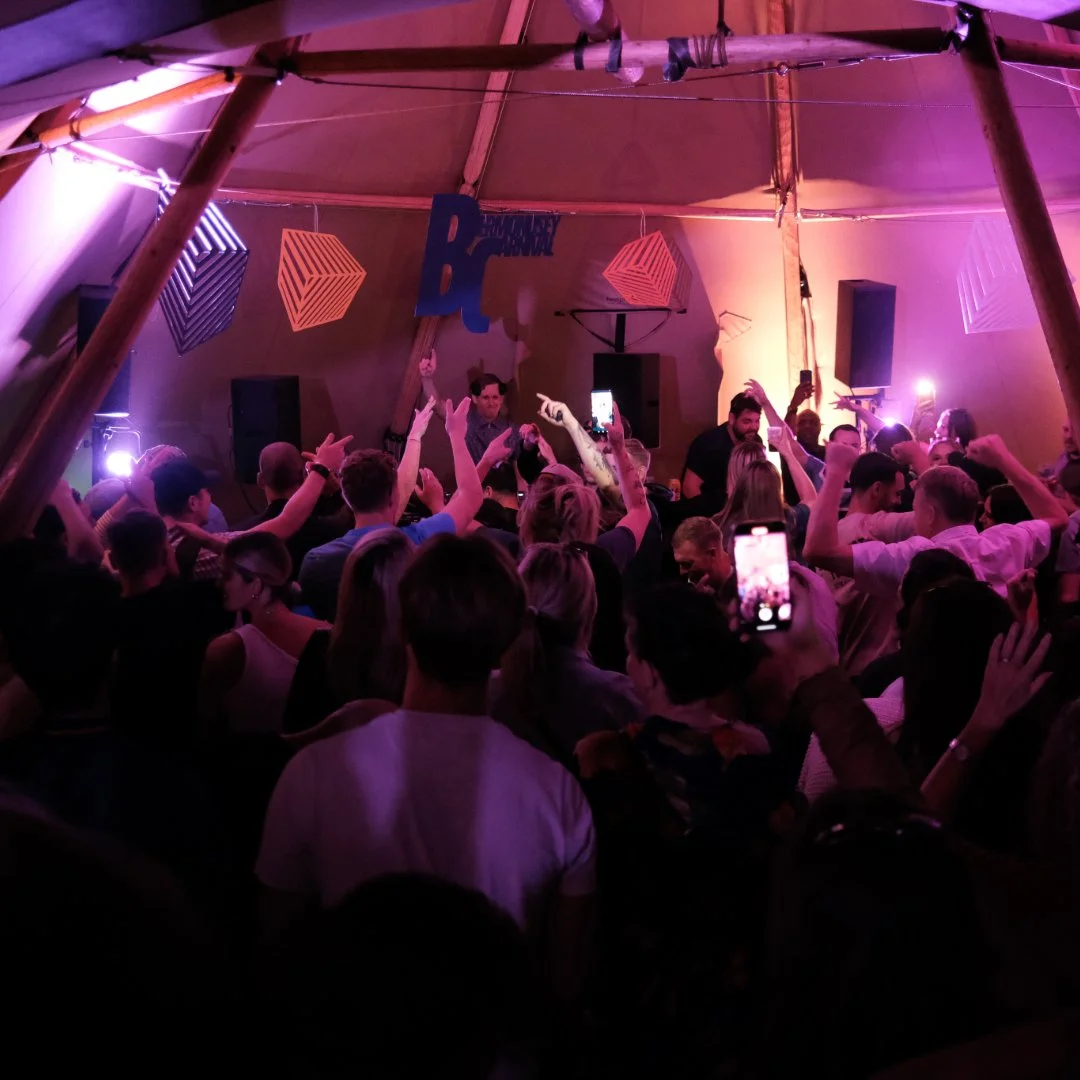 Crowd at a nightclub or party dancing with purple and pink lighting, with DJ and speakers in the background.