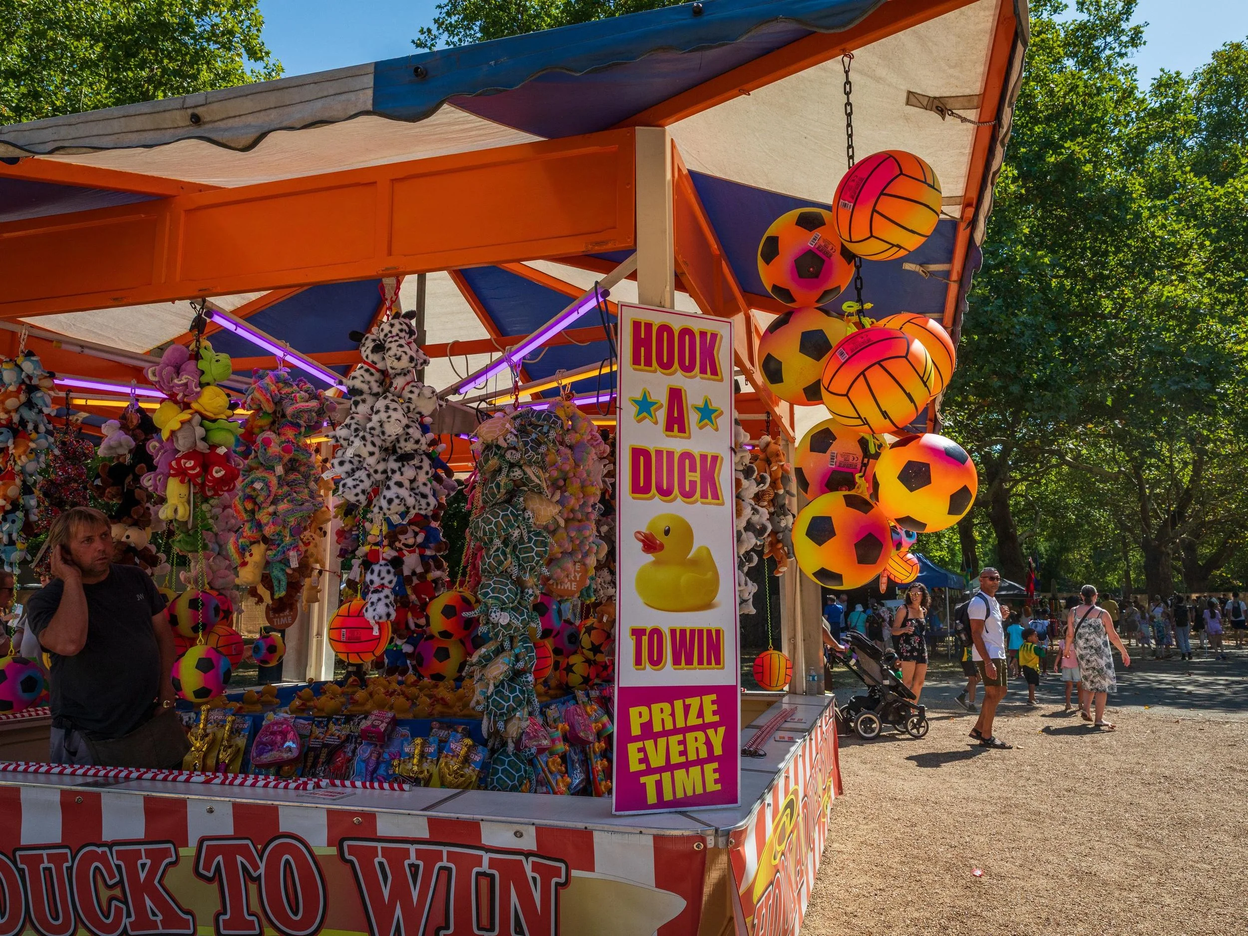 A carnival game booth with stuffed animal prizes hanging, colorful themed soccer and volleyball balls, and neon lights. People walk around in the background on a sunny day with leafy trees.