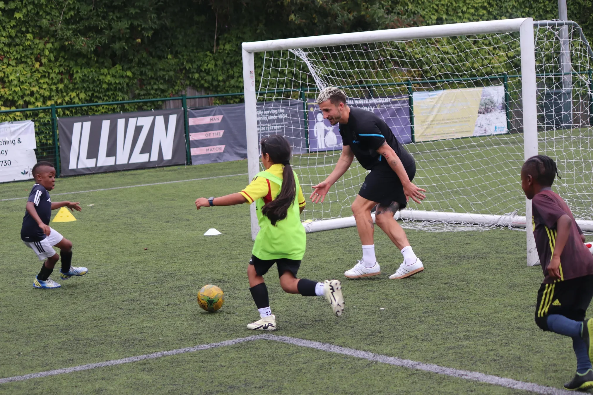 A man and children playing soccer on a field. The man is bending and pointing, wearing a black shirt and shorts; children are running around, one with a yellow jersey, another wearing a black and yellow uniform, and a boy in a navy shirt and white shorts. There is a goal net behind them and trees in the background.