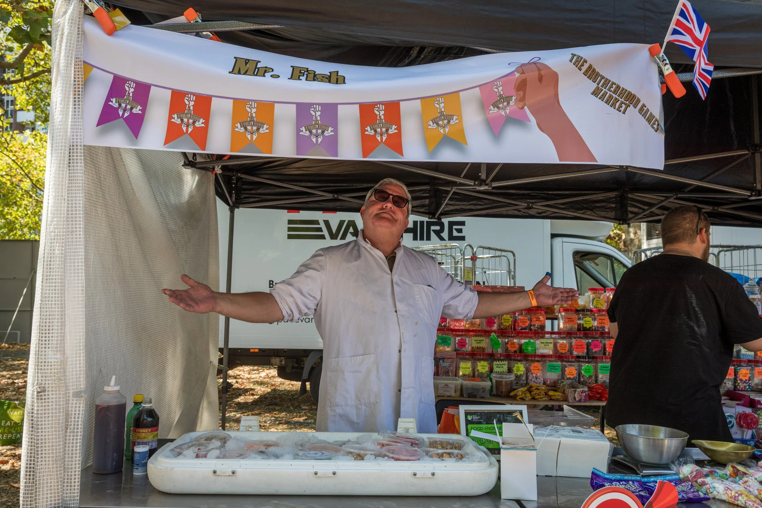 A man in a white apron with sunglasses standing behind a candy and snack stall at the Brothershood Candy Market, with his arms outstretched and a welcoming gesture, under a sign that reads "Mr. Fish" and "The Brotherhood Candy Market". The stall displays various packaged sweets and candies, with a group of people around, and trees in the background.