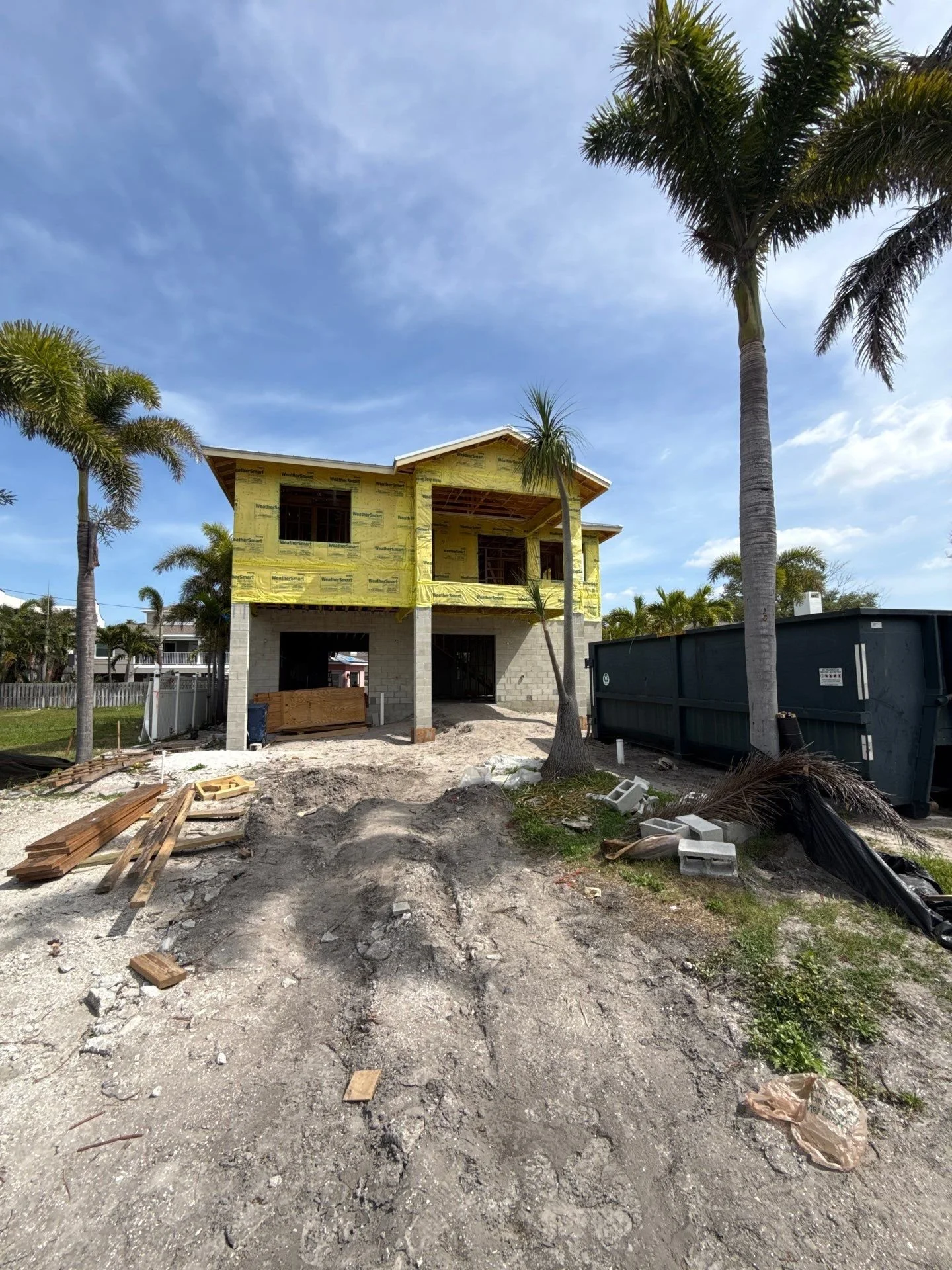 Construction site of a house with yellow insulation, palm trees, construction debris, wood planks, a Dumpster, and a partly cloudy sky.