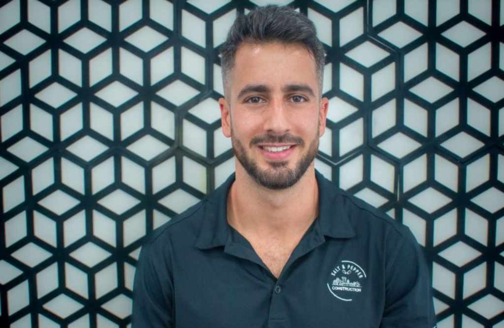 A smiling man with short dark hair, a beard, and light skin, wearing a dark blue polo shirt with a construction logo, standing in front of a geometric patterned wall.