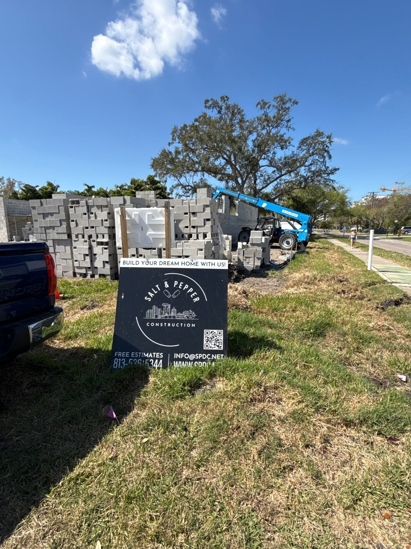 Construction site with stacked concrete blocks, a small excavator in the background, and a sign that reads "Build your dream home with us! Salt & Pepper Construction" with contact information, on a sunny day with a blue sky and trees.
