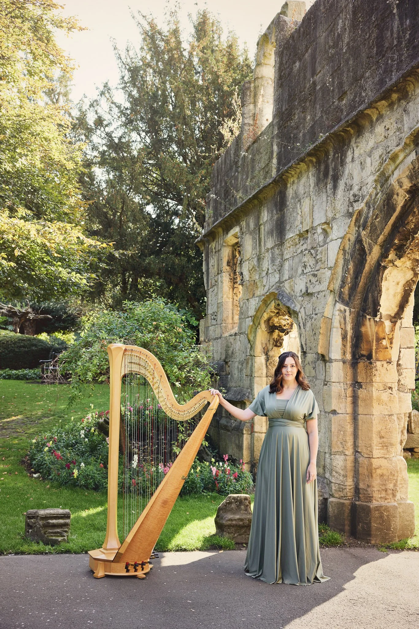 A woman in a long, flowing, gray-green gown stands outdoors in front of an old stone building, next to a pedal harp. The setting is lush with greenery and colorful flowers, and there are trees in the background.