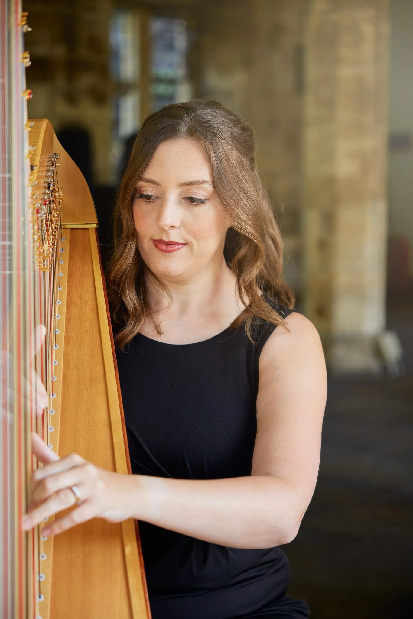 A woman with brown hair styled in loose waves, wearing a black sleeveless top, playing a wooden harp in a room with large windows and brick walls.