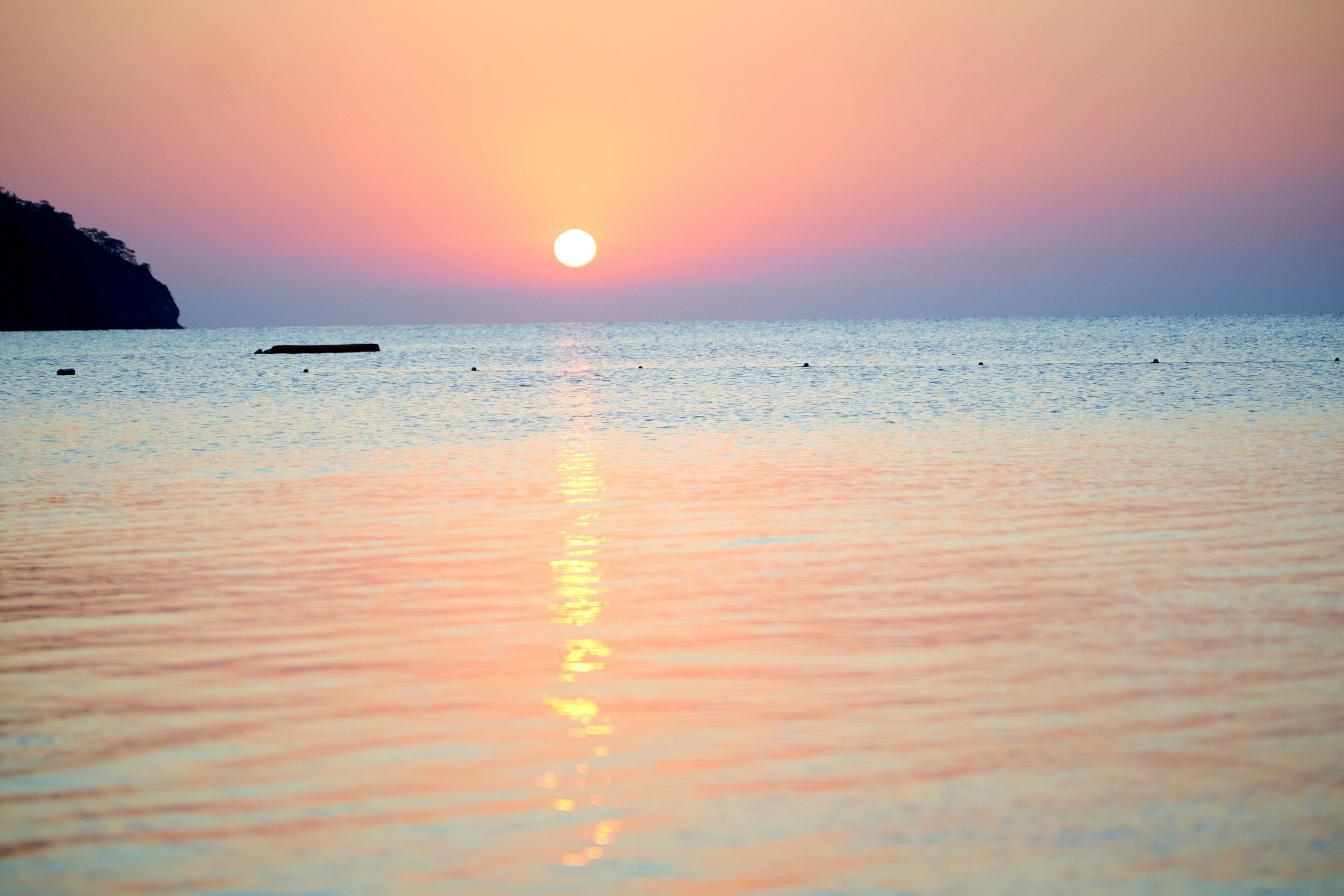 Sunrise or sunset over the ocean with a pink and orange sky, a silhouette of a cliff on the left, and a boat floating on the water.