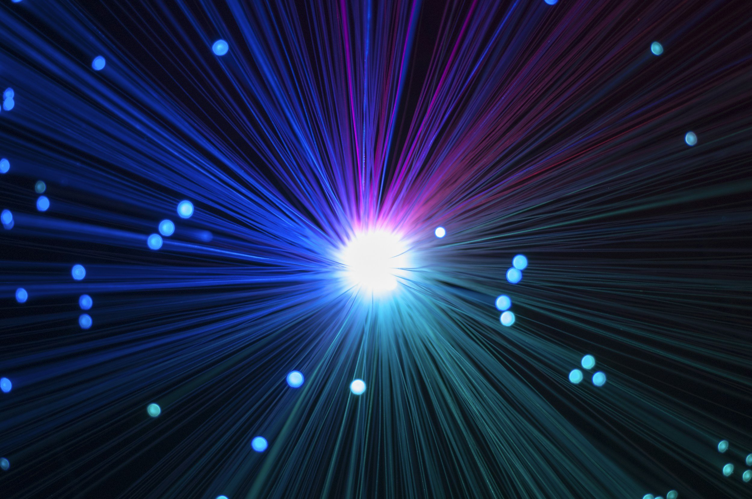 Abstract digital image with bright multicolored rays emanating from a central white light point, with small glowing blue dots scattered throughout.